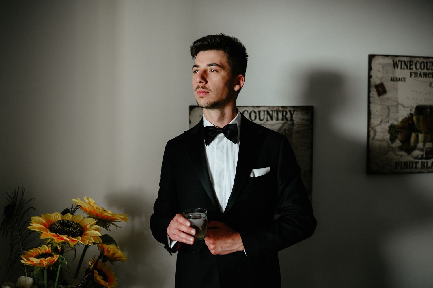Groom in black tuxedo with bow tie holding a glass, standing near sunflowers and vintage wall art.