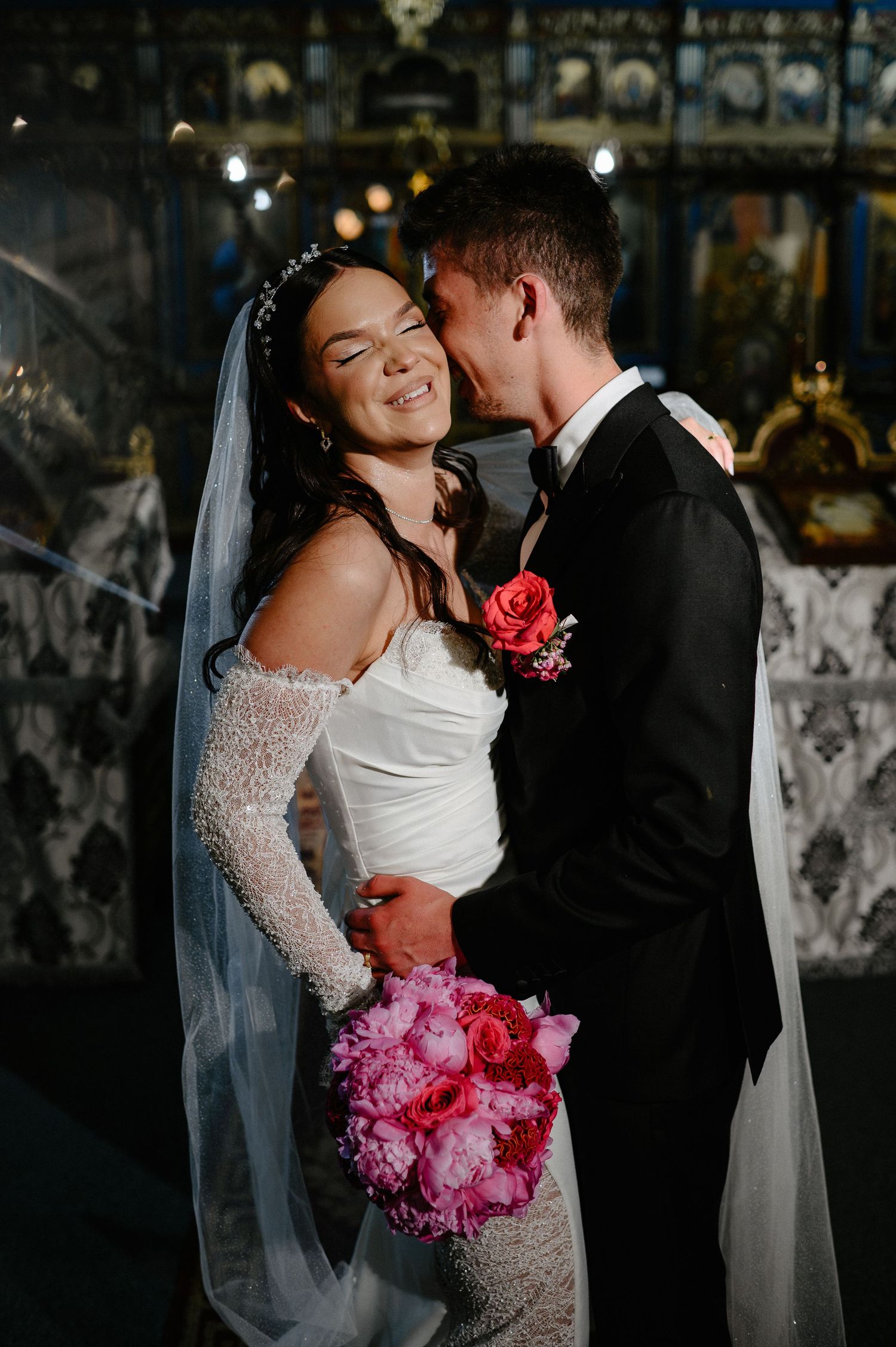 Groom tenderly kisses smiling bride wearing veil and lace gloves holding pink peony bouquet in church.