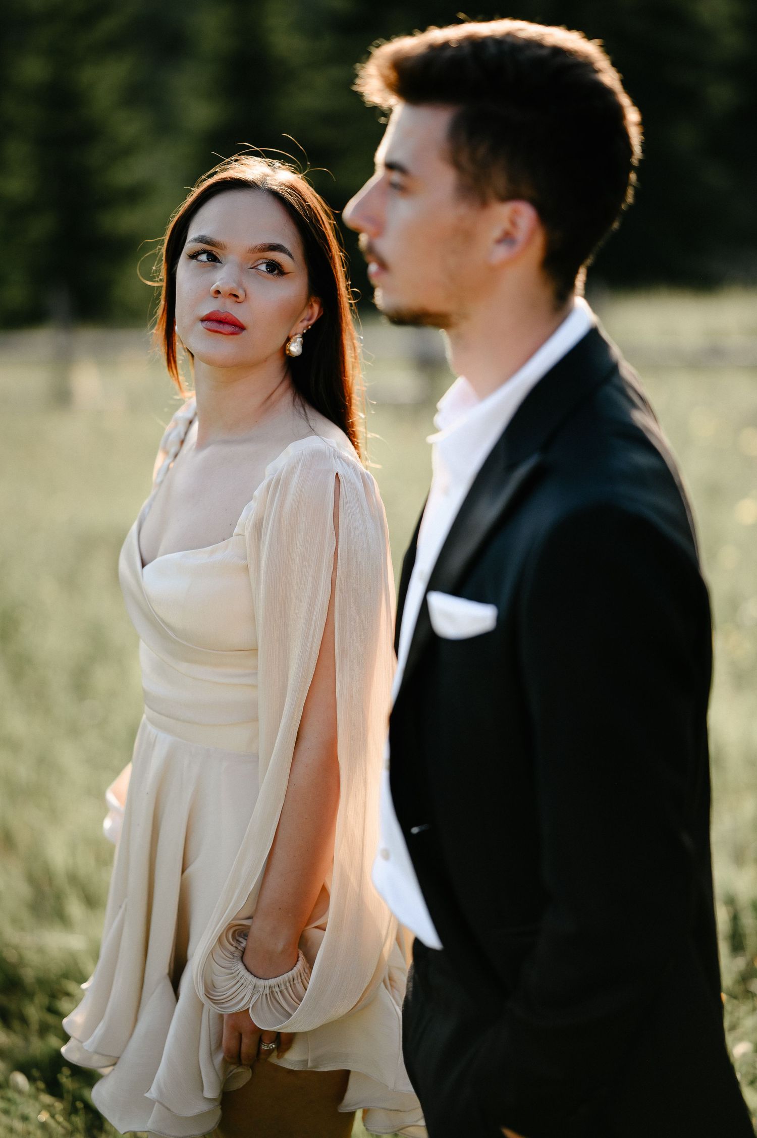 Bride in flowing cream dress with cape sleeves stands beside groom in black suit in a meadow.
