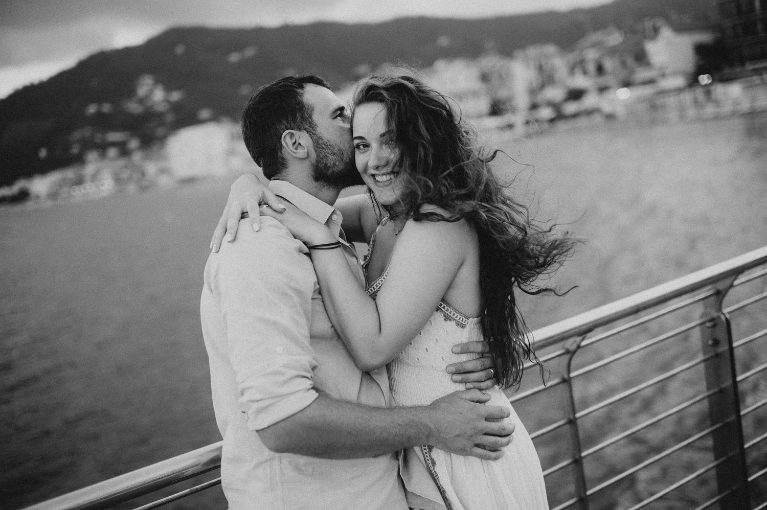 Black and white romantic couple embracing on a waterfront railing with a scenic coastal town behind them.