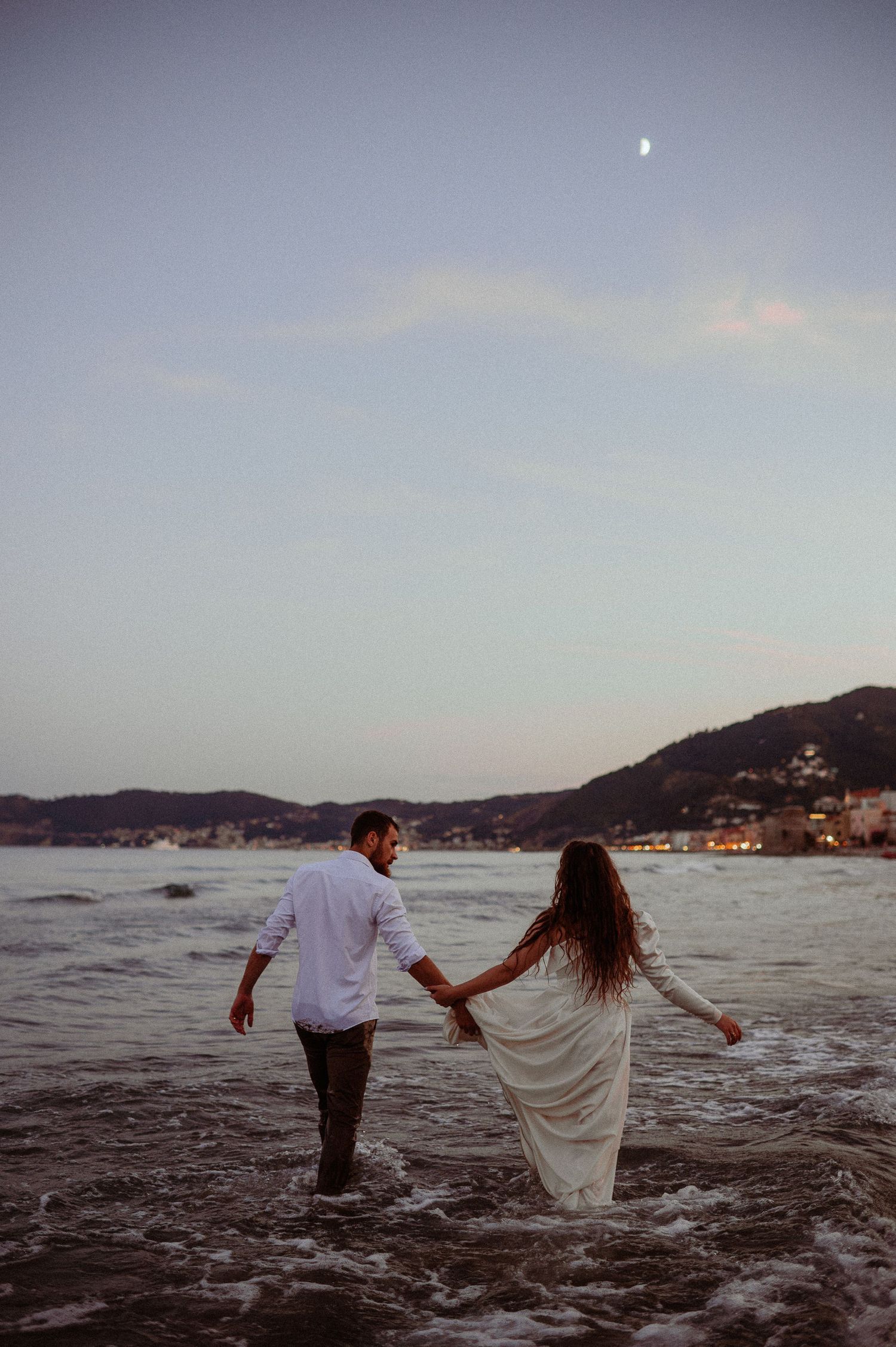 Couple holding hands walking into ocean waves at twilight with crescent moon and illuminated coastal town behind.