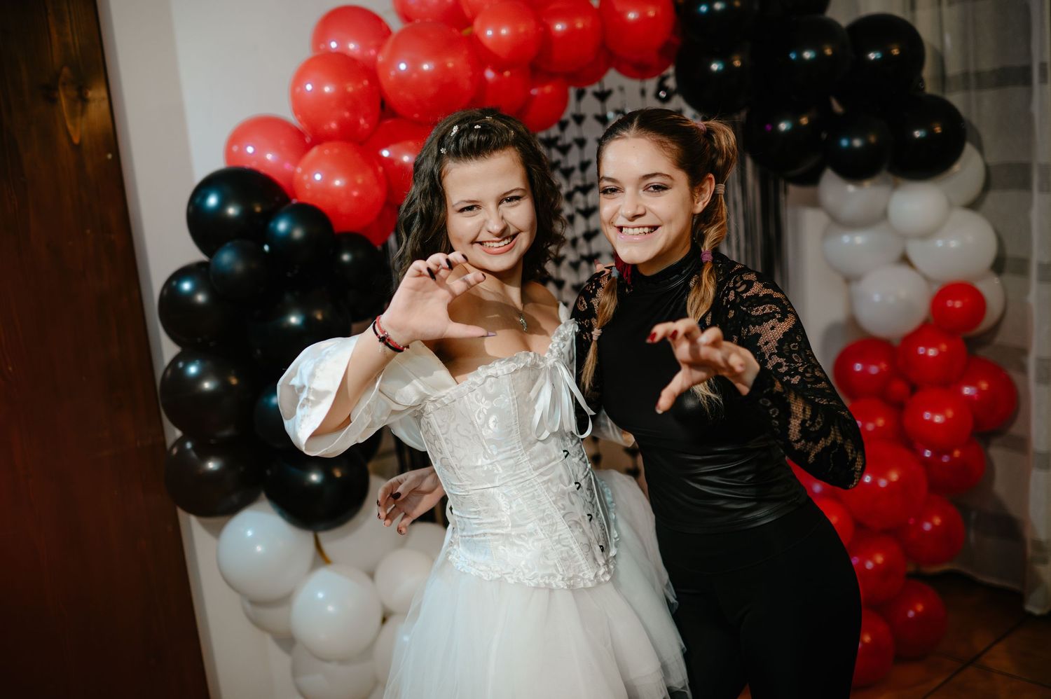 Two young women in Halloween costumes smile and pose before colorful balloon arch decoration.