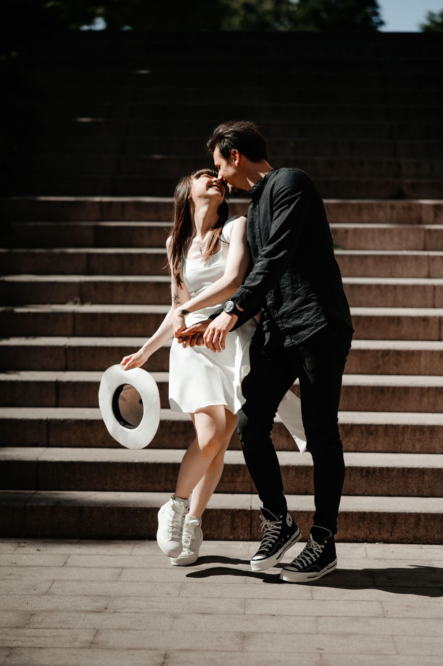 Happy couple laughing and embracing on sunlit stone steps, woman in white dress holding a white hat.