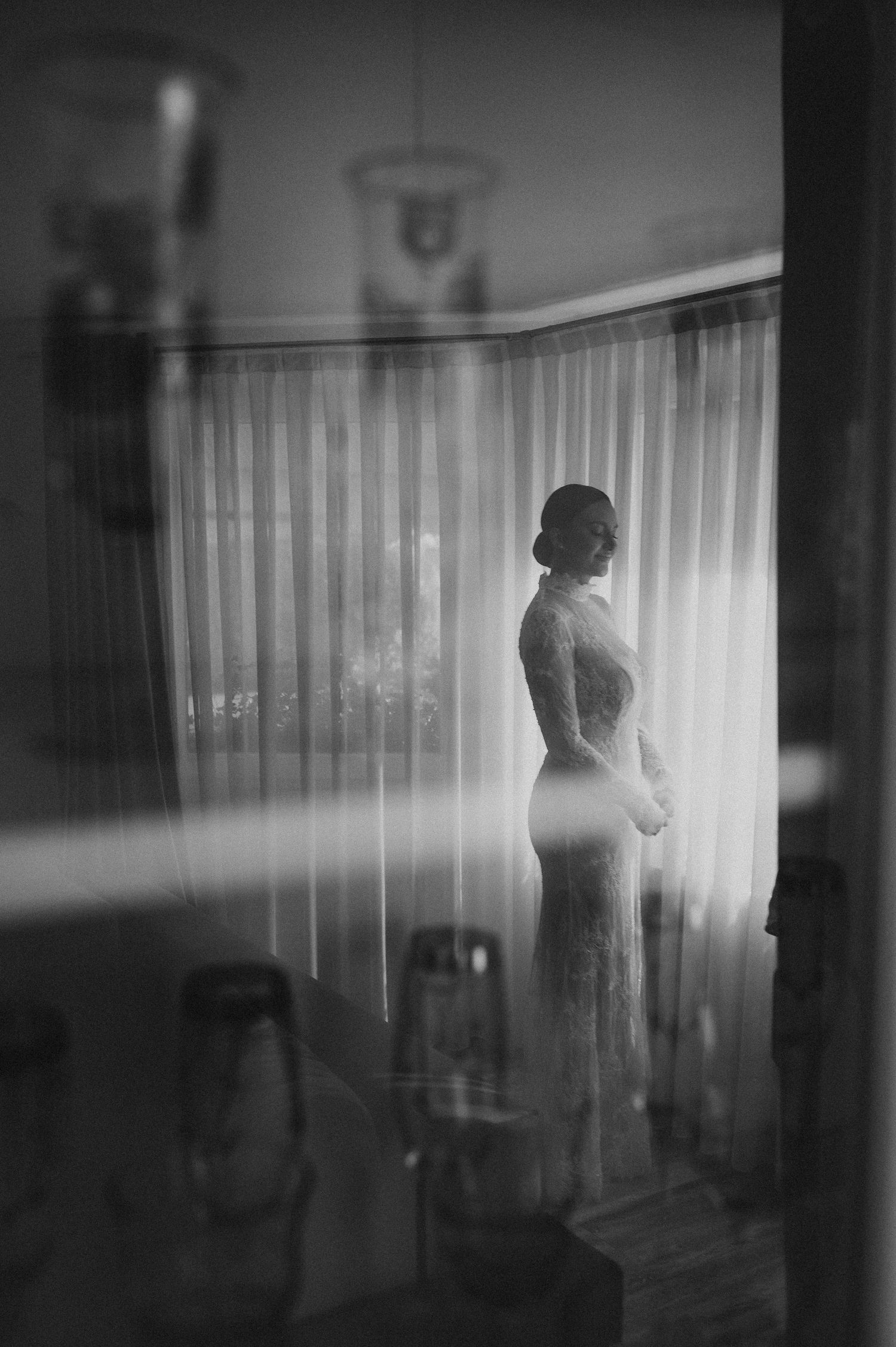 Bride in lace wedding dress silhouetted against sheer curtains, photographed through a reflective surface in black and white.
