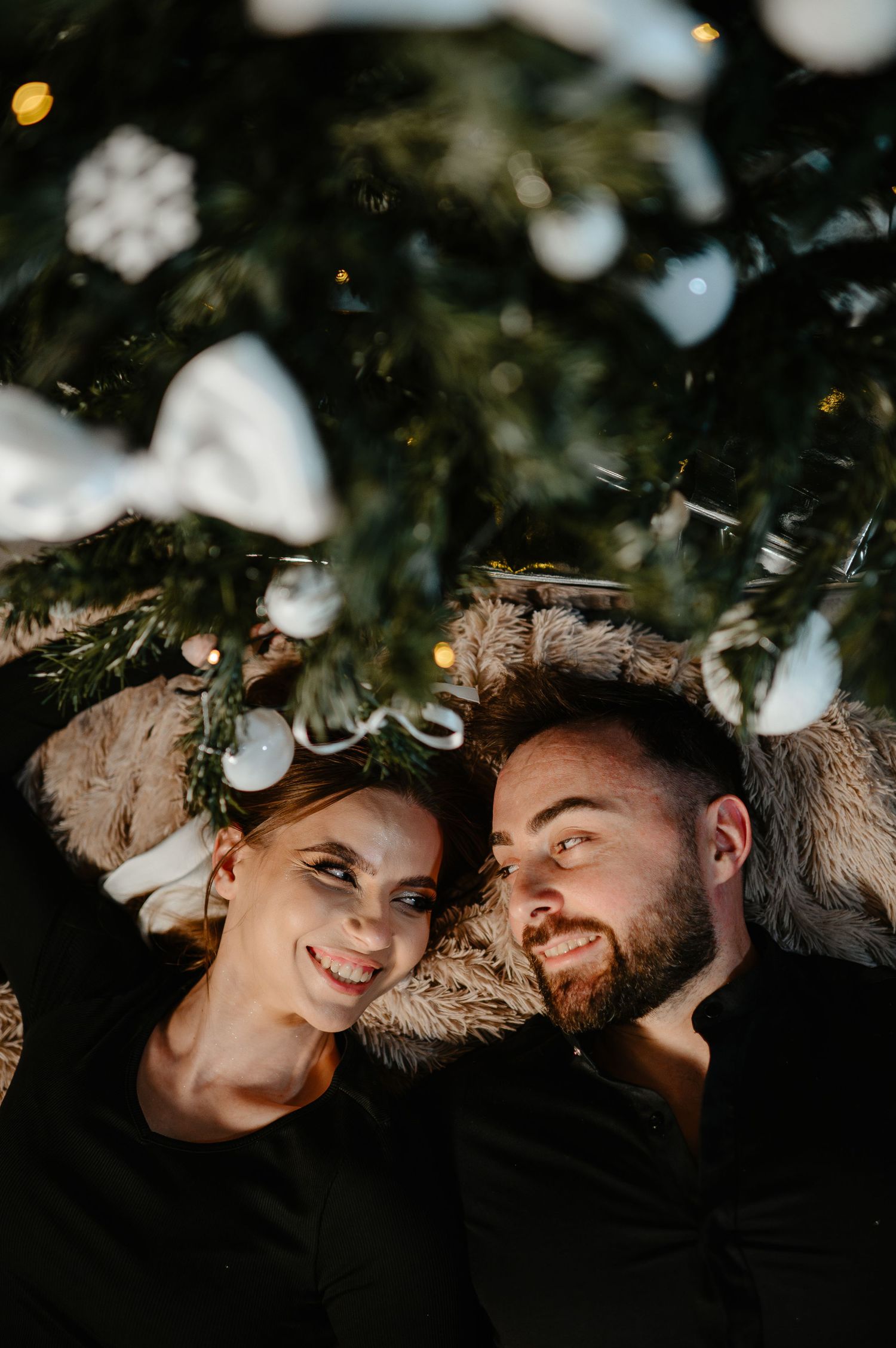 Couple lying under a decorated Christmas tree smiling at each other on a fur blanket.