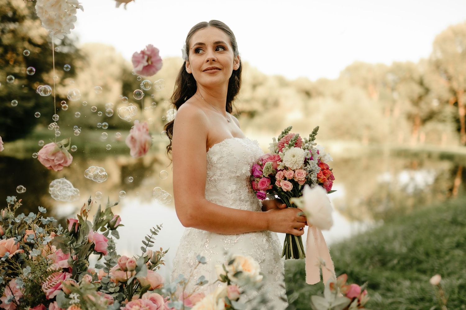Elegant bride in strapless wedding dress holding pink bouquet with bubbles floating in golden outdoor setting.