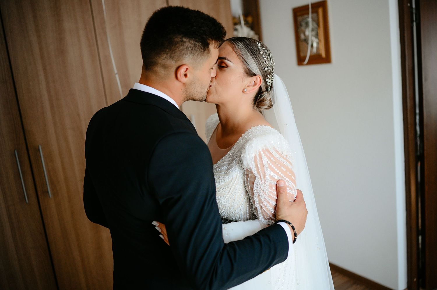 Bride and groom sharing romantic kiss, groom in dark suit, bride in beaded lace wedding gown with veil.