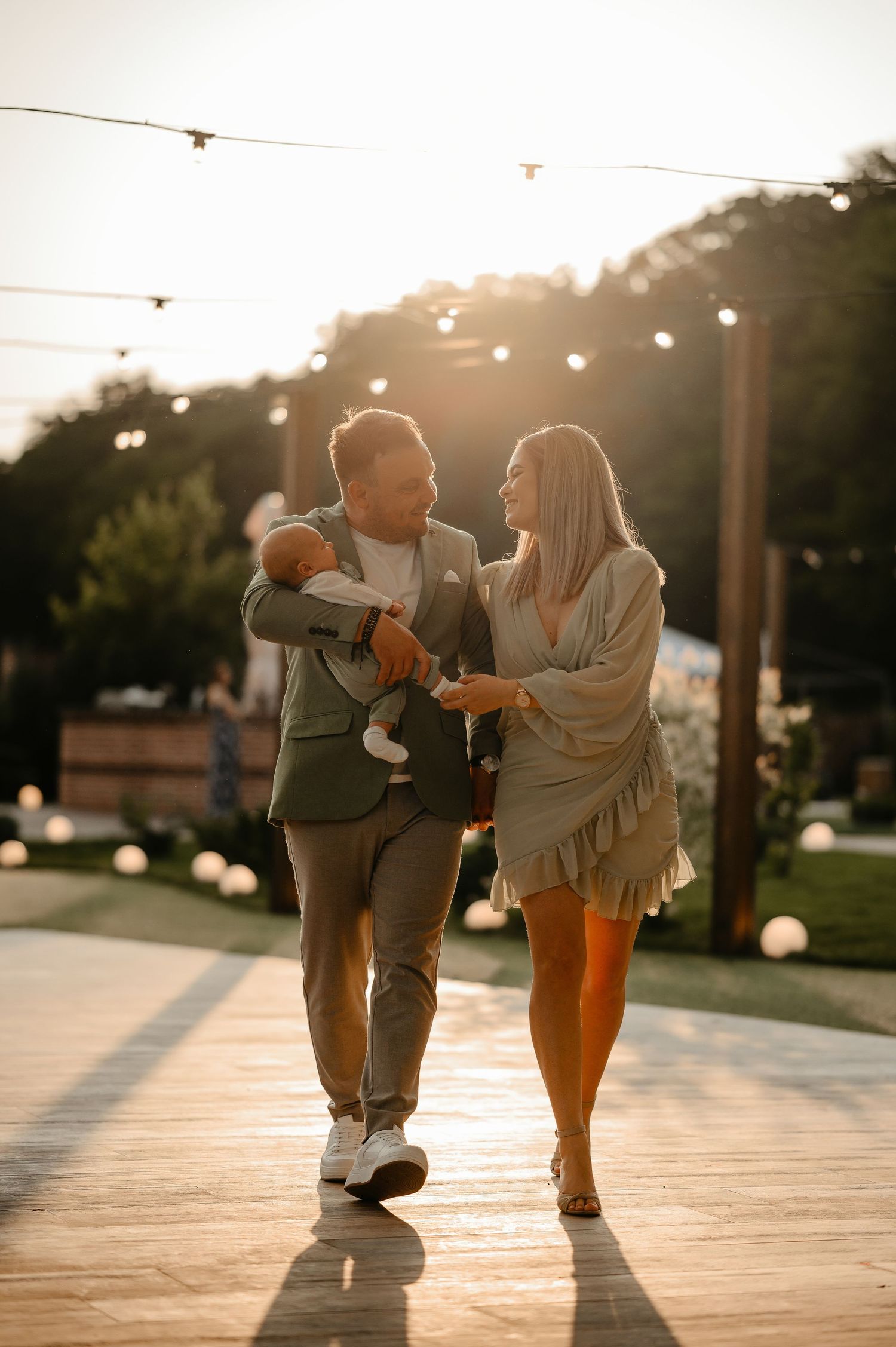 A couple walks outdoors at golden hour, man holding a newborn baby while exchanging a loving glance with the woman.