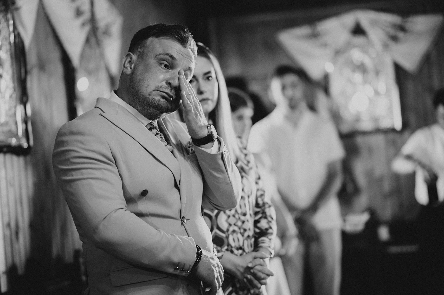 Emotional couple in beige attire wipe tears during a moving religious christening ceremony.
