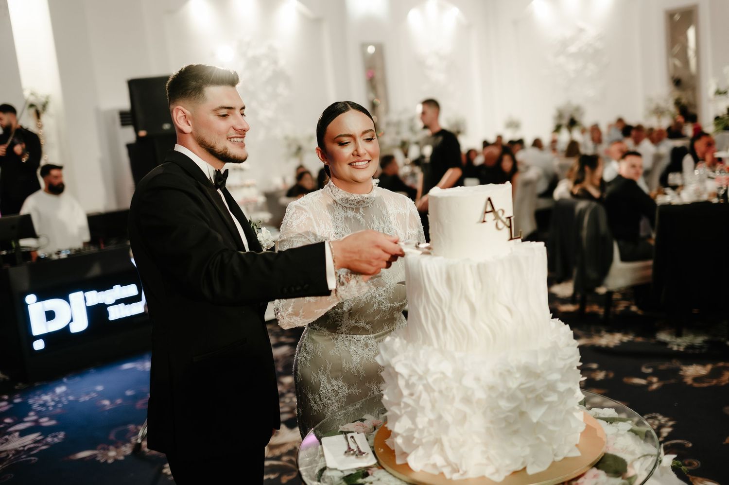 Smiling bride and groom cutting their elegant white ruffled wedding cake with gold monogram topper.