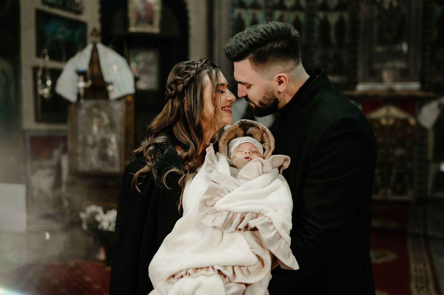 Happy parents in black outfits sharing tender moment with newborn at baptism ceremony.