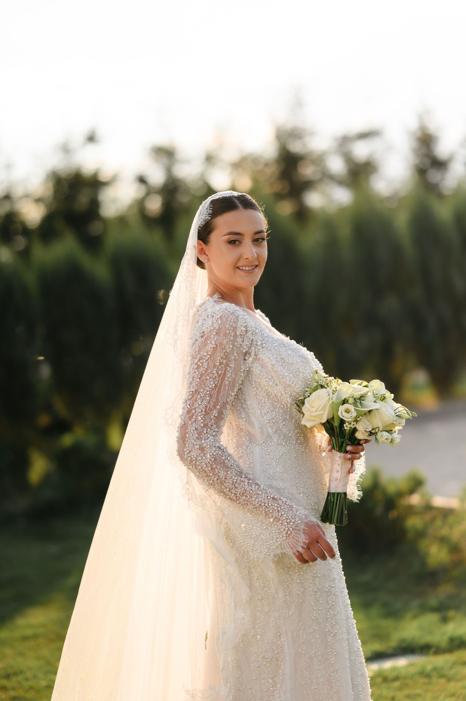 Elegant bride in a beaded long-sleeve white gown and veil holds a white bouquet outdoors.