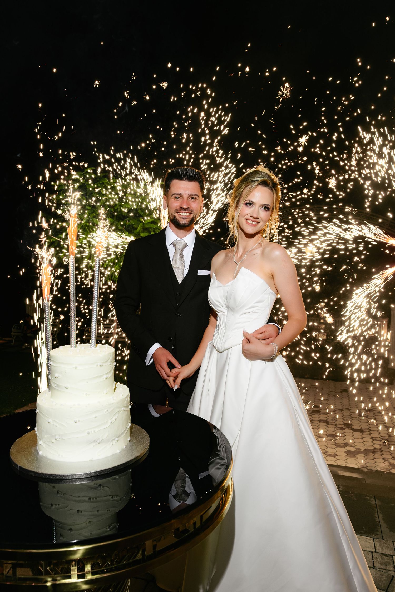 Bride and groom smile beside white wedding cake with sparklers and fireworks display at night.