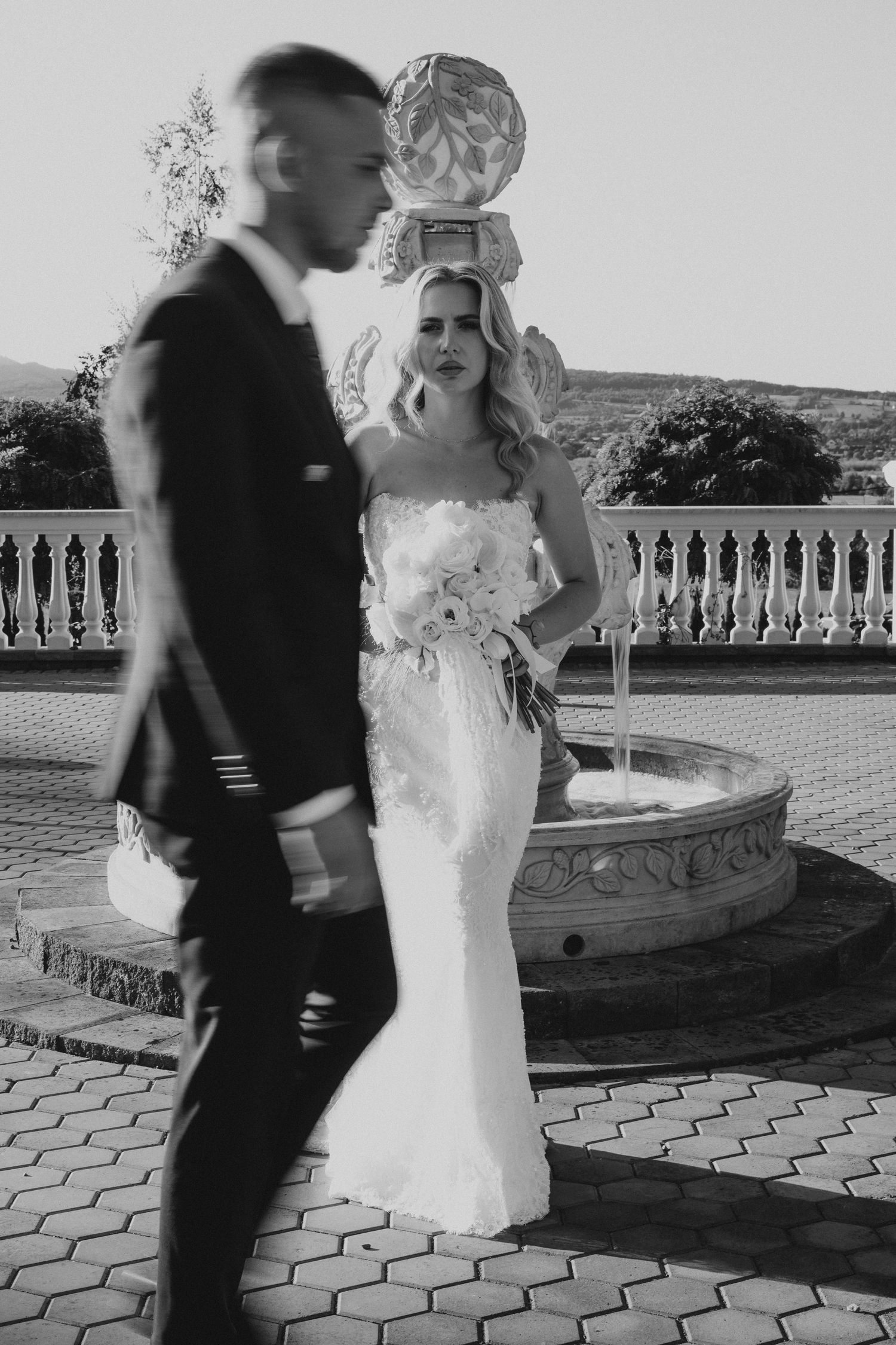 Black and white wedding photo of bride in white gown holding bouquet by fountain, groom in suit walking past.