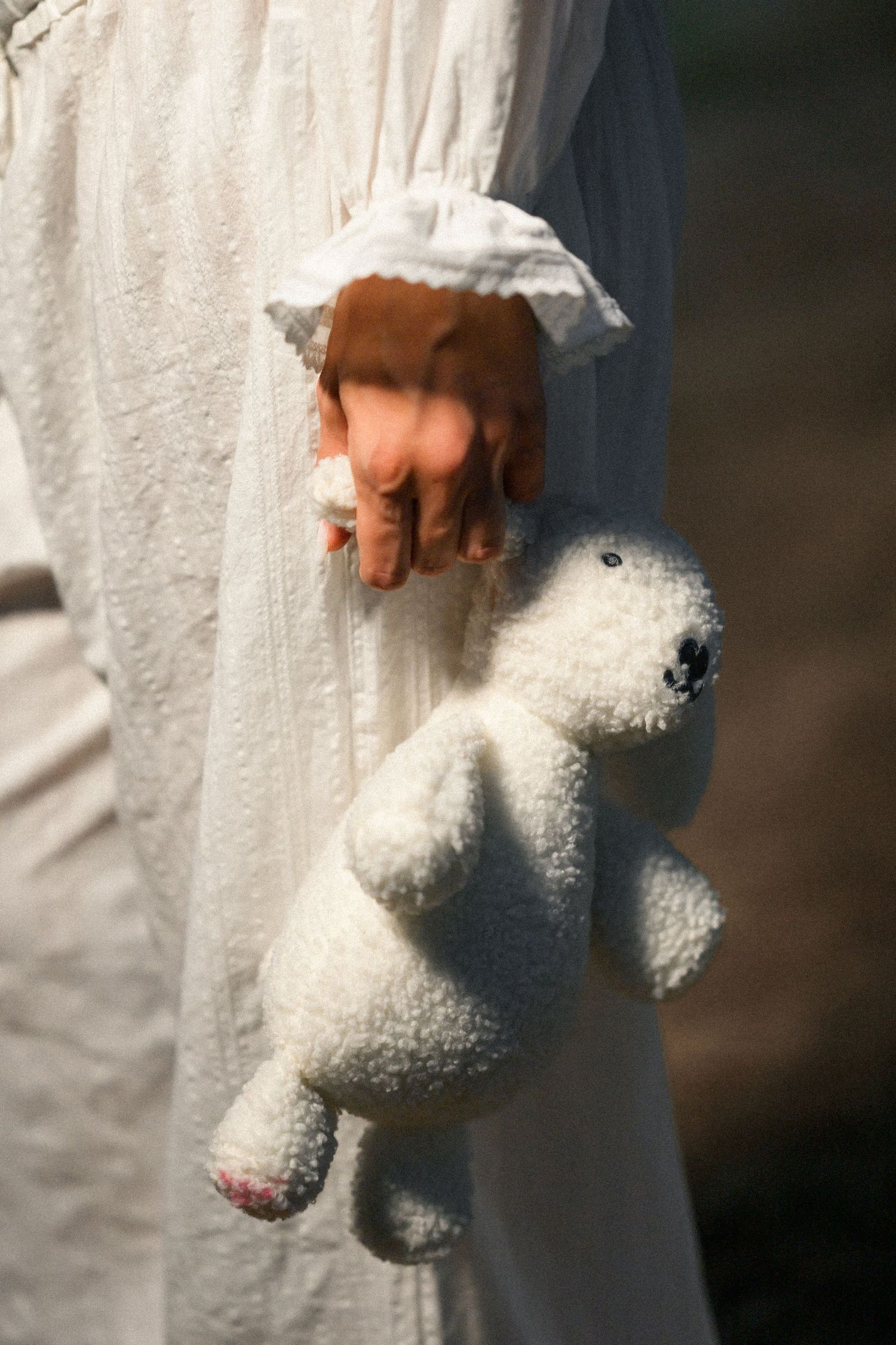 Child in white dress holding a small fluffy white teddy bear toy, captured in soft natural light.