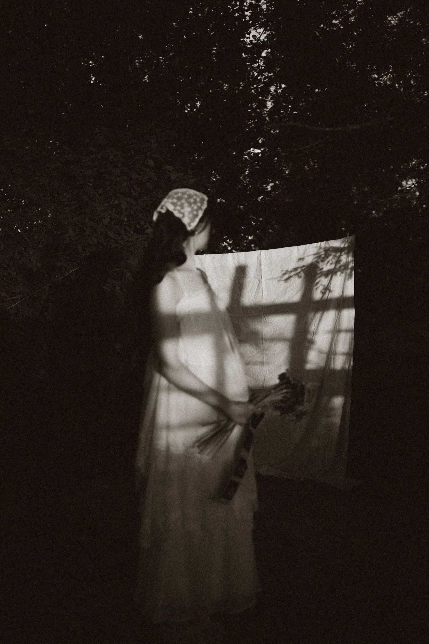 Black and white photo of a woman in white dress hanging laundry outdoors in a dark, moody forest setting.