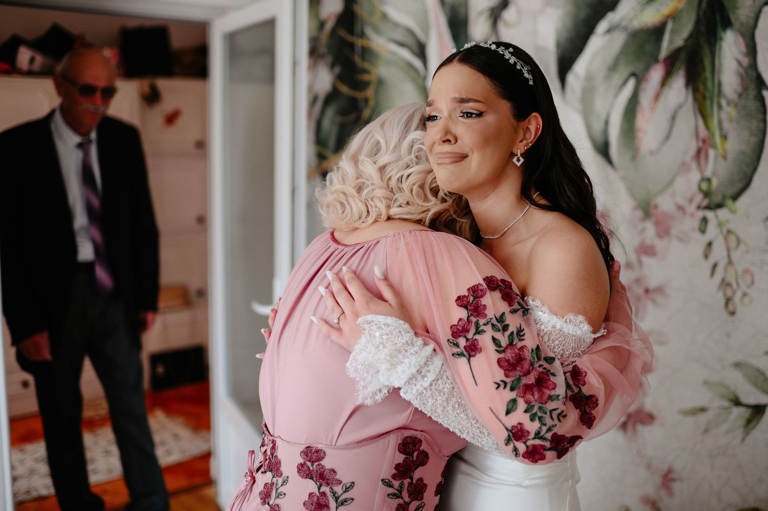 Emotional bride in white wedding dress sharing heartfelt hug with older woman in pink floral blouse.
