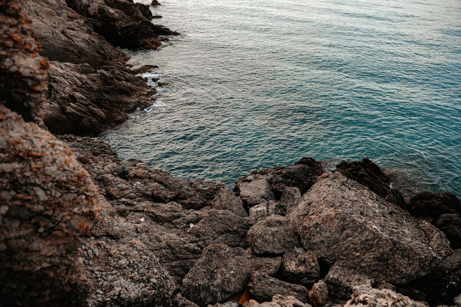 Rocky coastal cliffside overlooking calm turquoise Mediterranean sea waters at dusk.