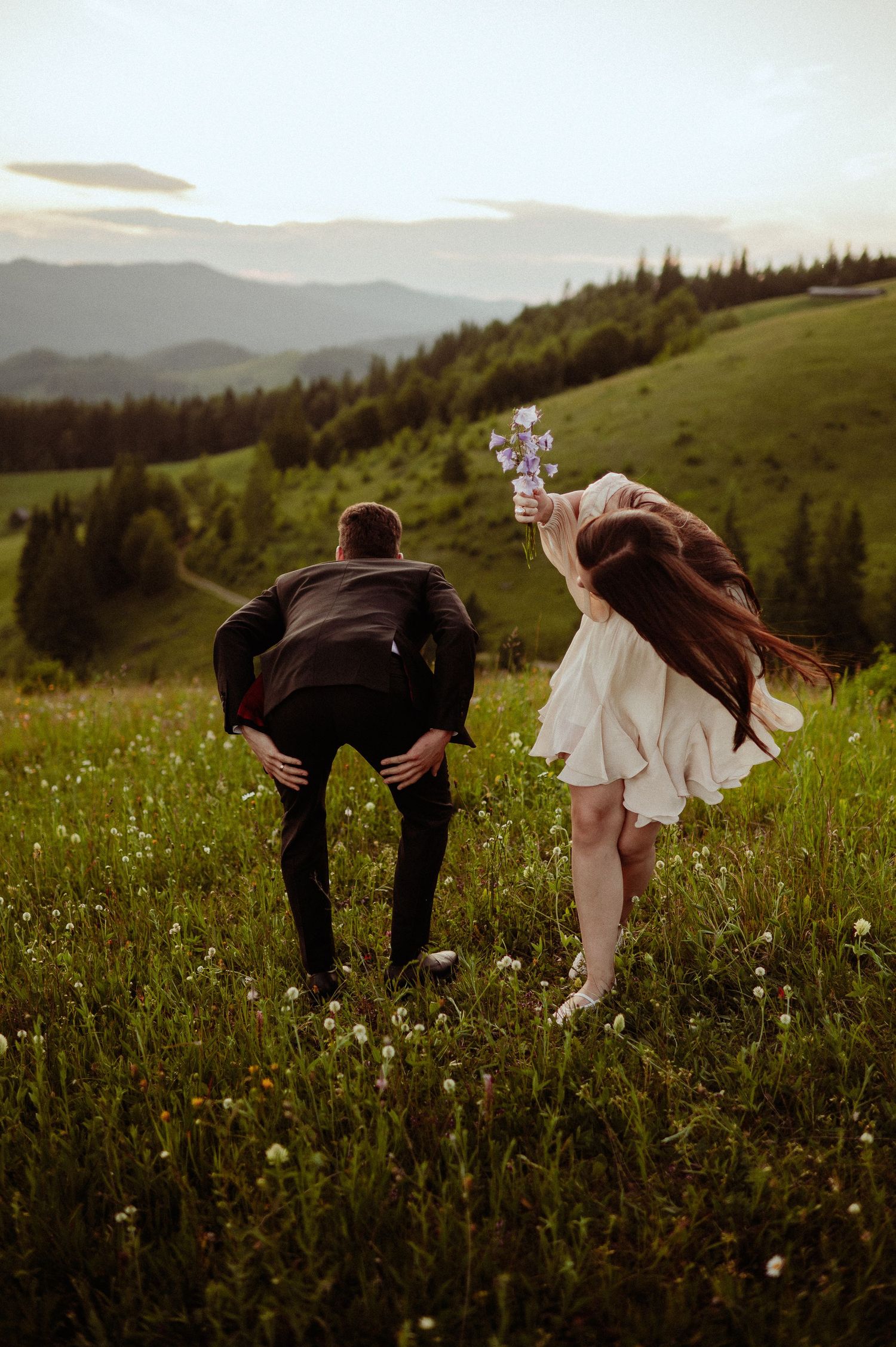 A playful couple laughs in a green mountain meadow, woman holding a pinwheel in the breeze.