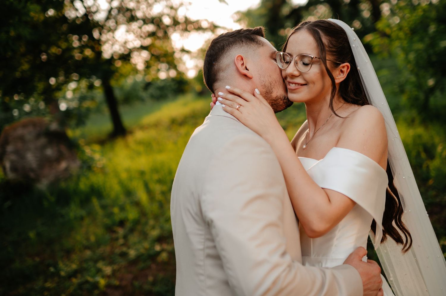Groom kissing bride wearing glasses and veil in sunlit garden during wedding photo session.