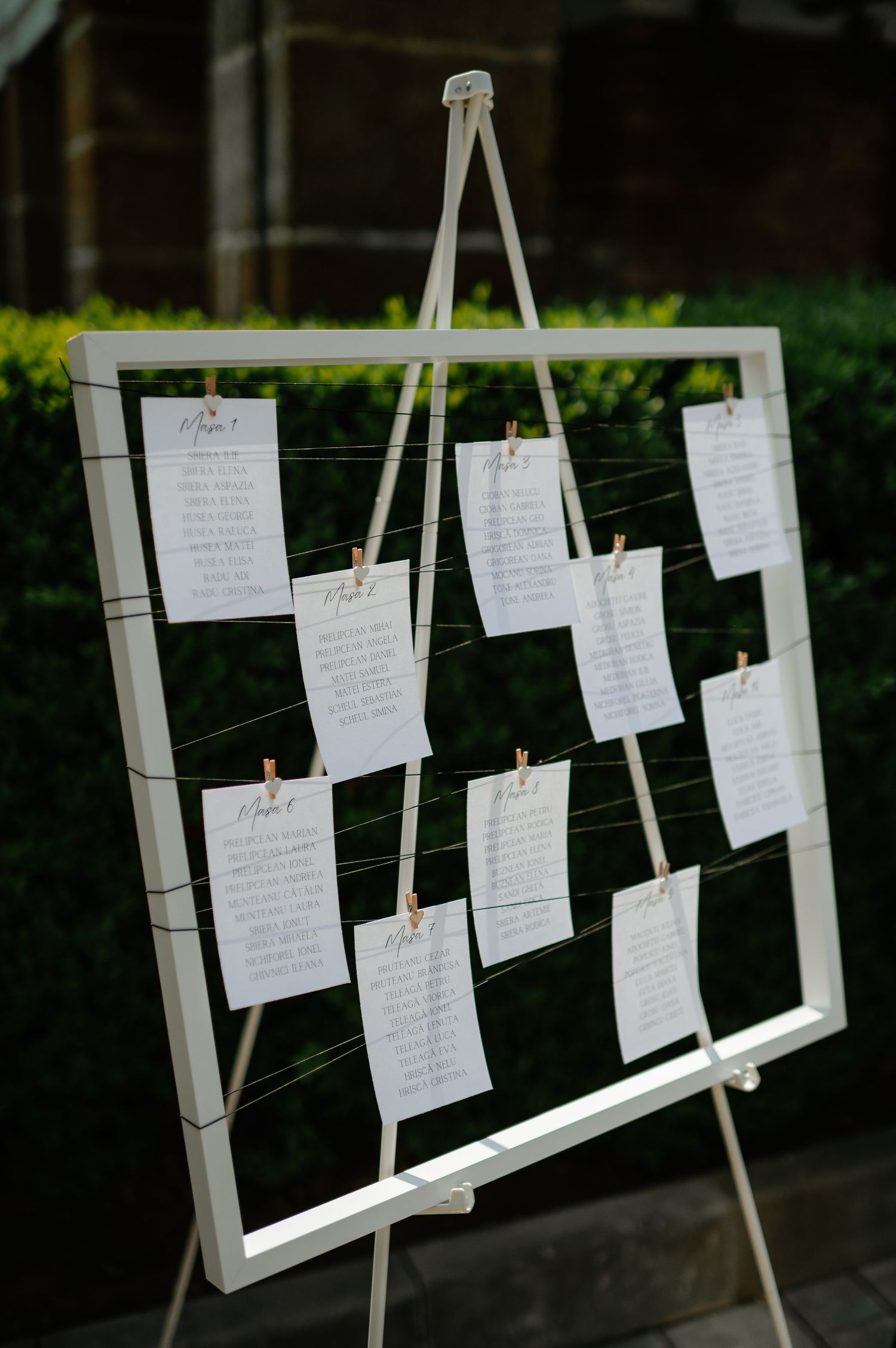 White wooden easel seating chart with handwritten guest name cards attached by clothespins at wedding.