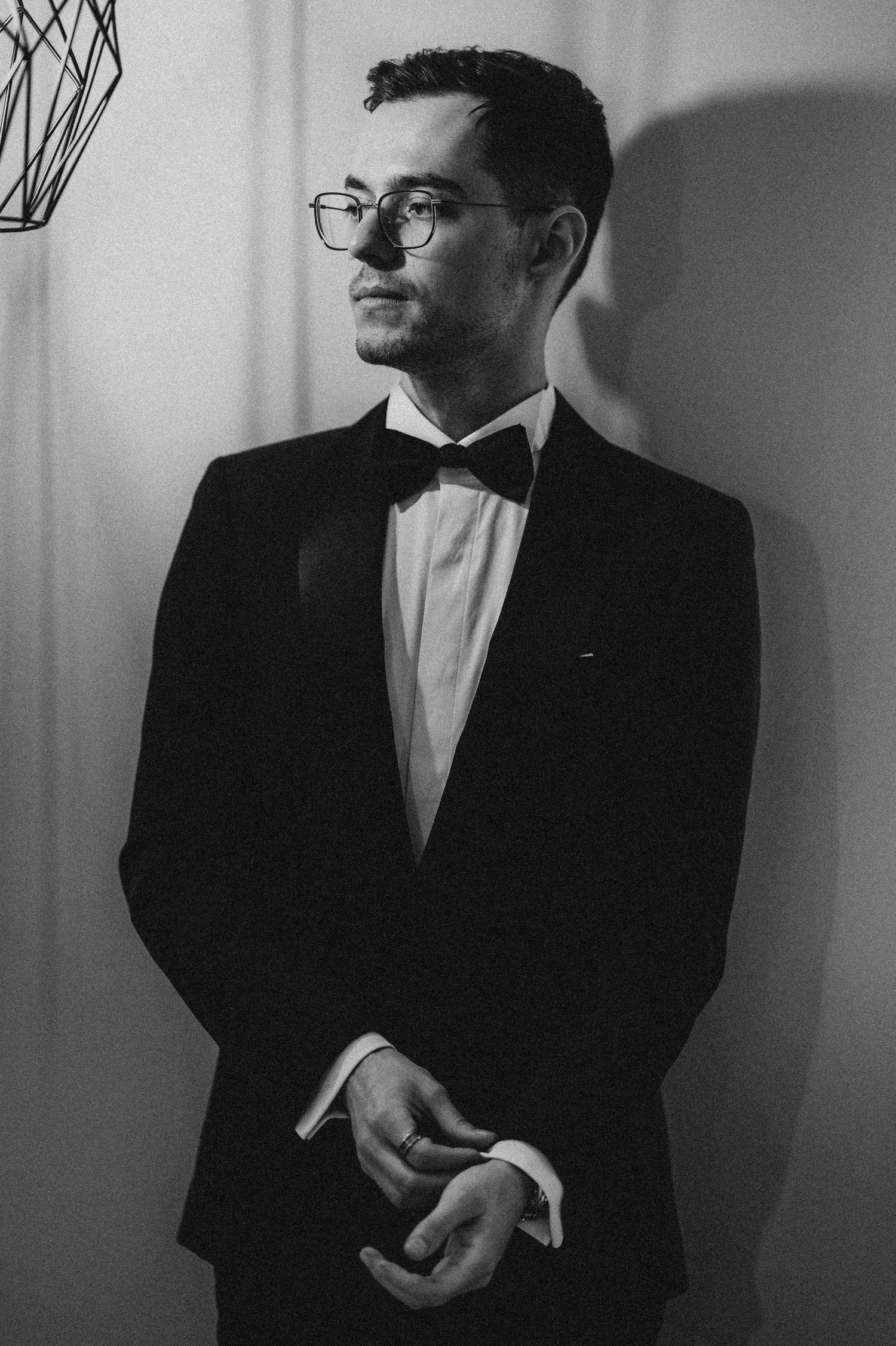 Black and white portrait of a man in a tuxedo with bow tie and glasses, posing elegantly indoors.