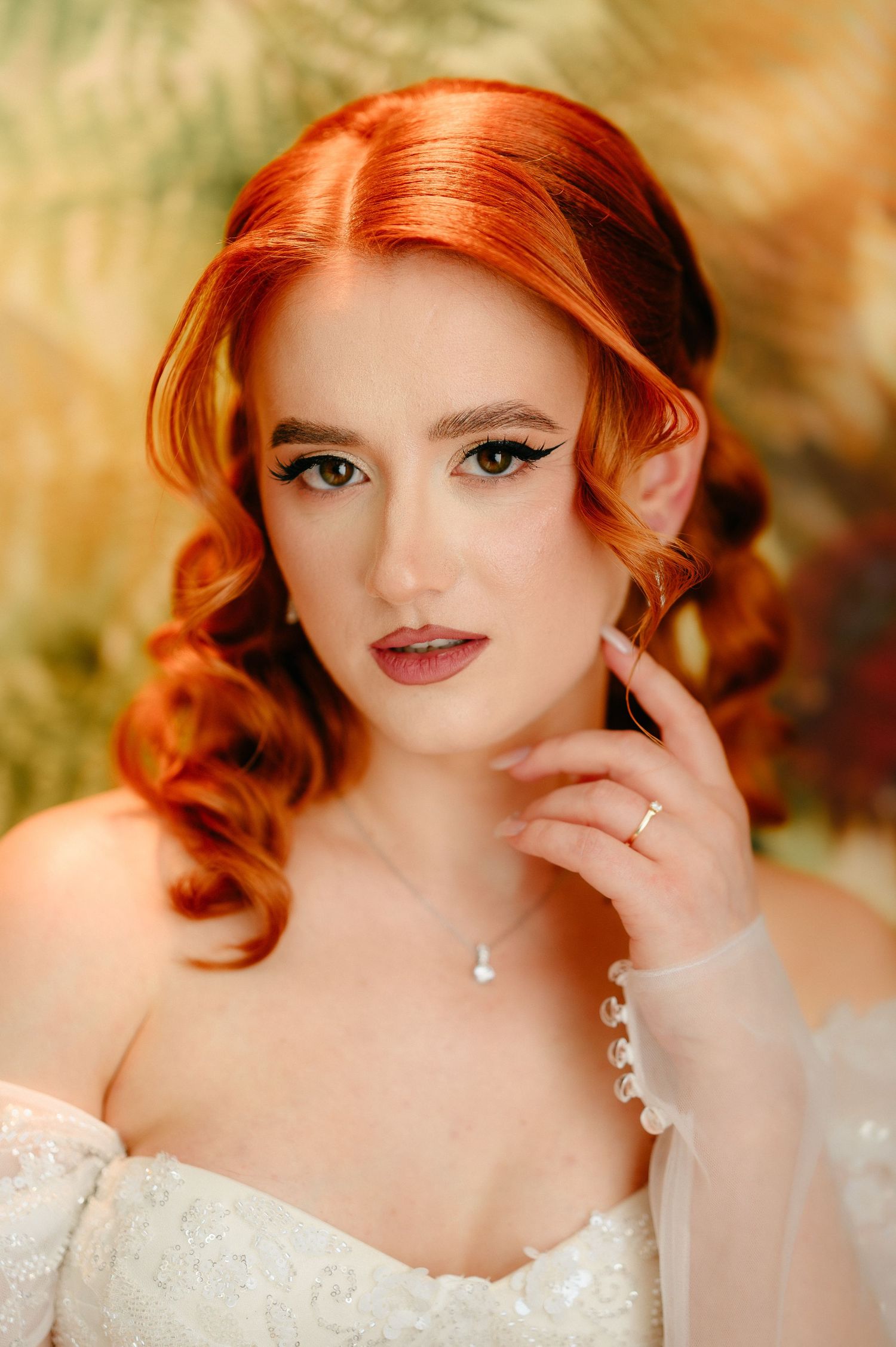 Elegant bride with red hair, cat-eye makeup, pearl necklace, and lace dress poses gracefully against a golden floral backdrop.