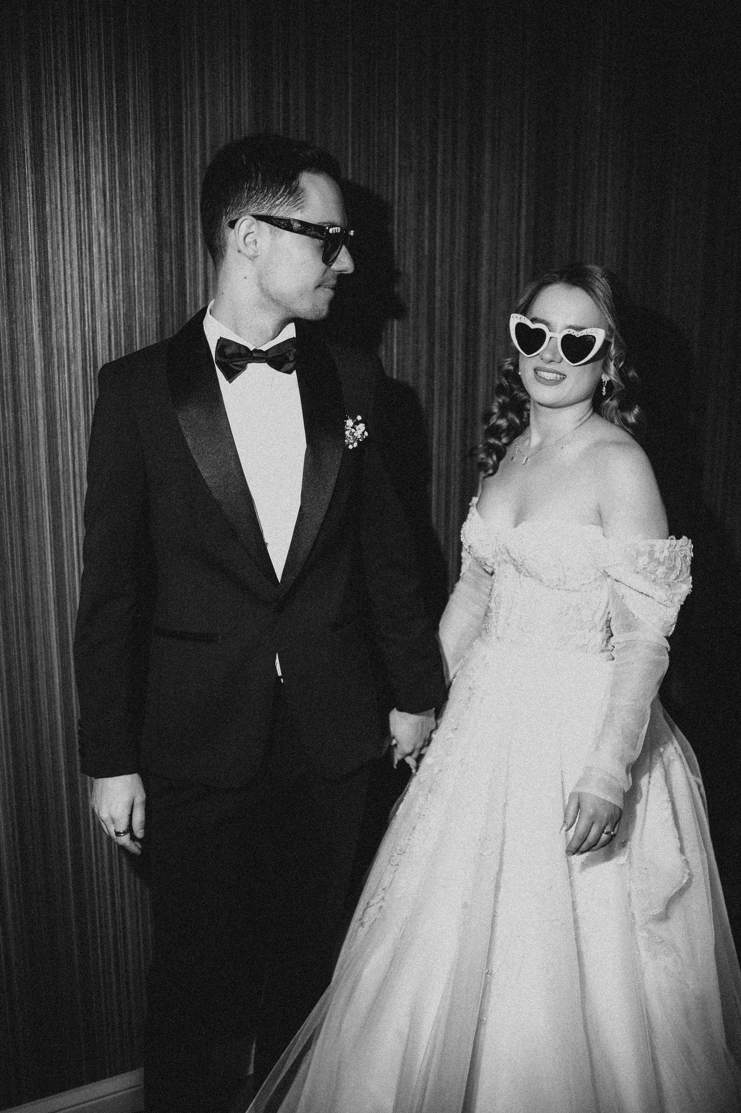 Black and white photo of a bride and groom holding hands, both wearing sunglasses, she in a ballgown, he in a tuxedo.