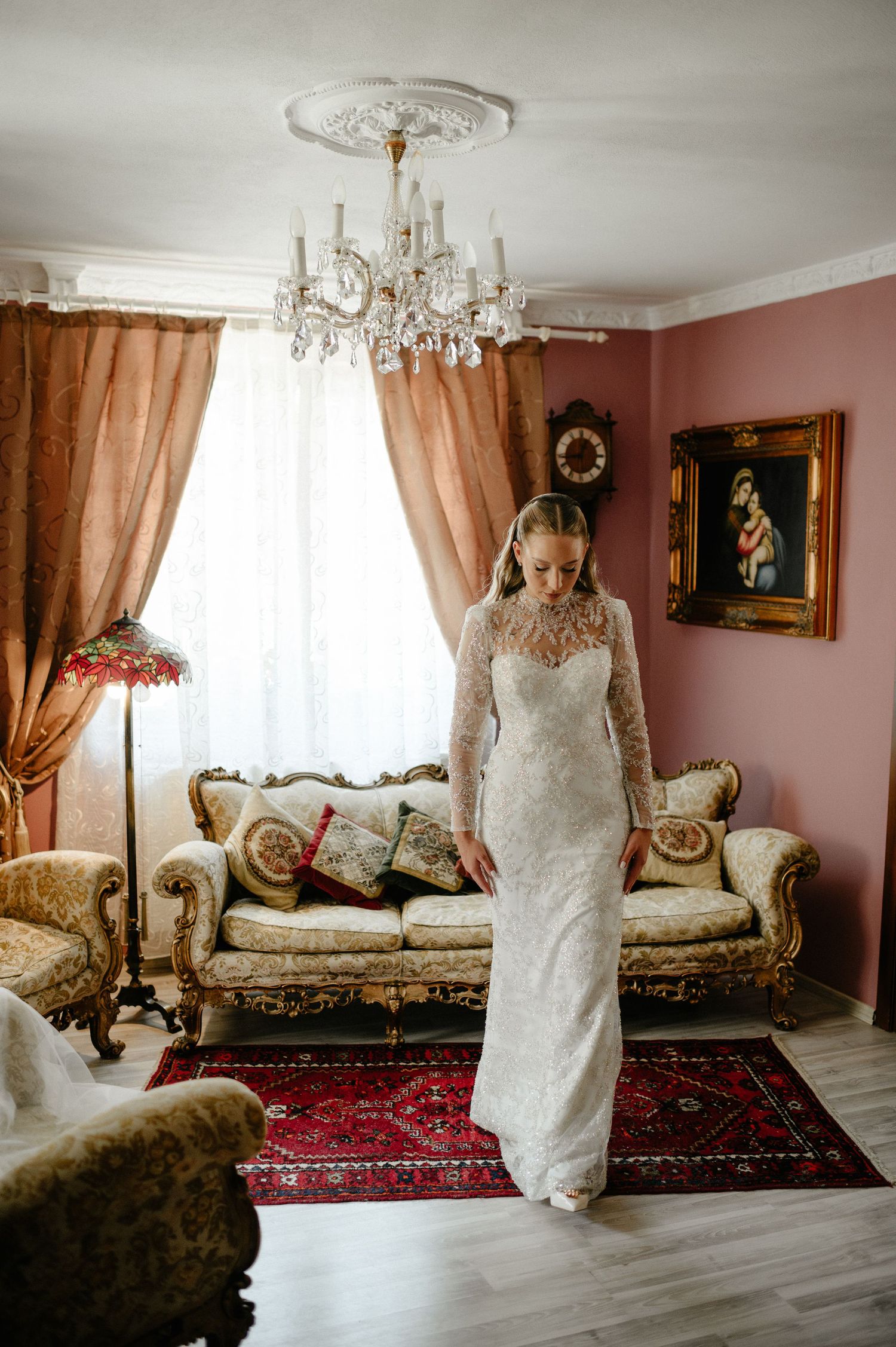 Elegant bride in long-sleeve lace wedding gown standing in vintage-styled room with crystal chandelier.