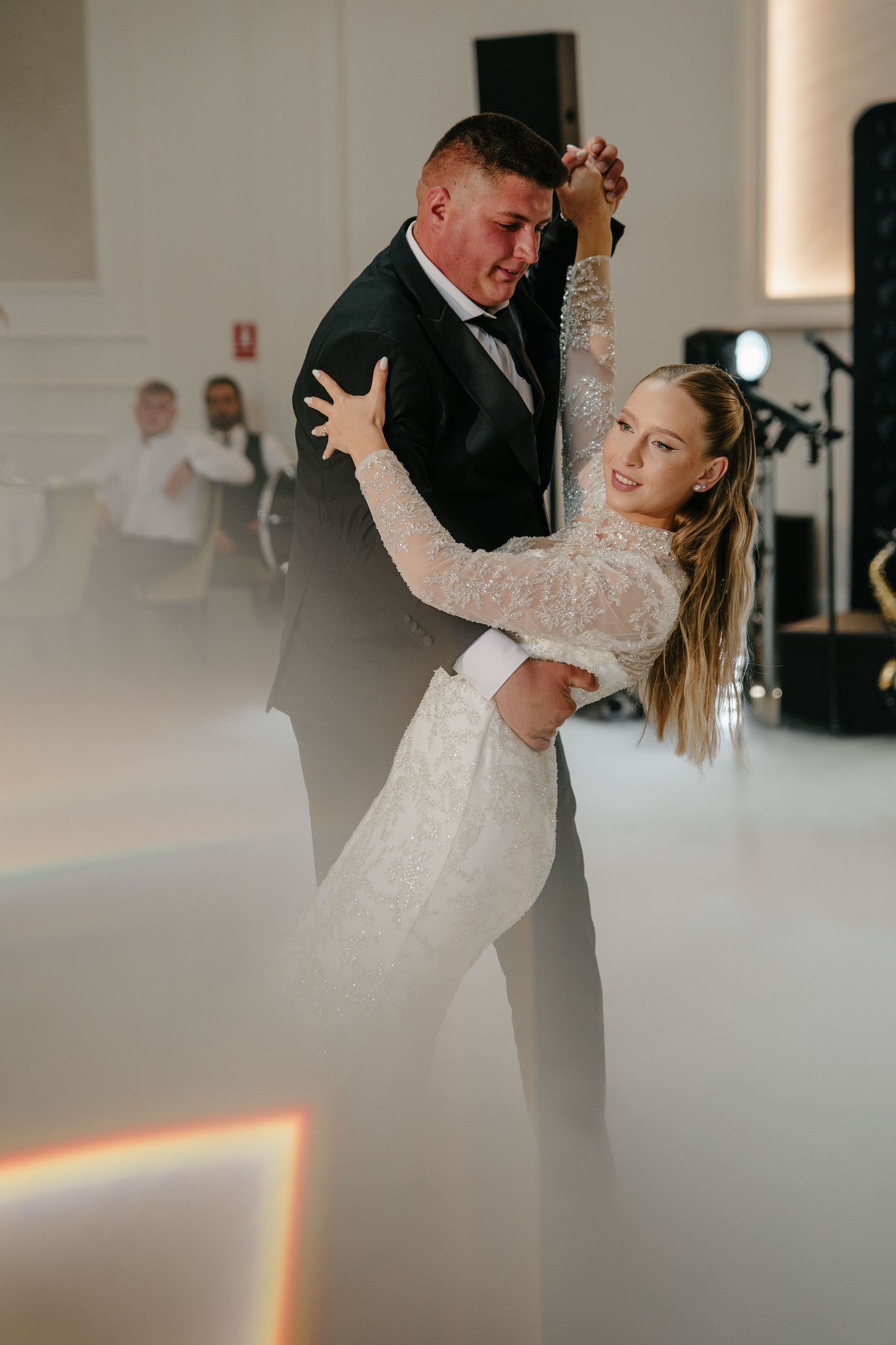 Bride and groom sharing romantic first dance surrounded by atmospheric fog on white dance floor.