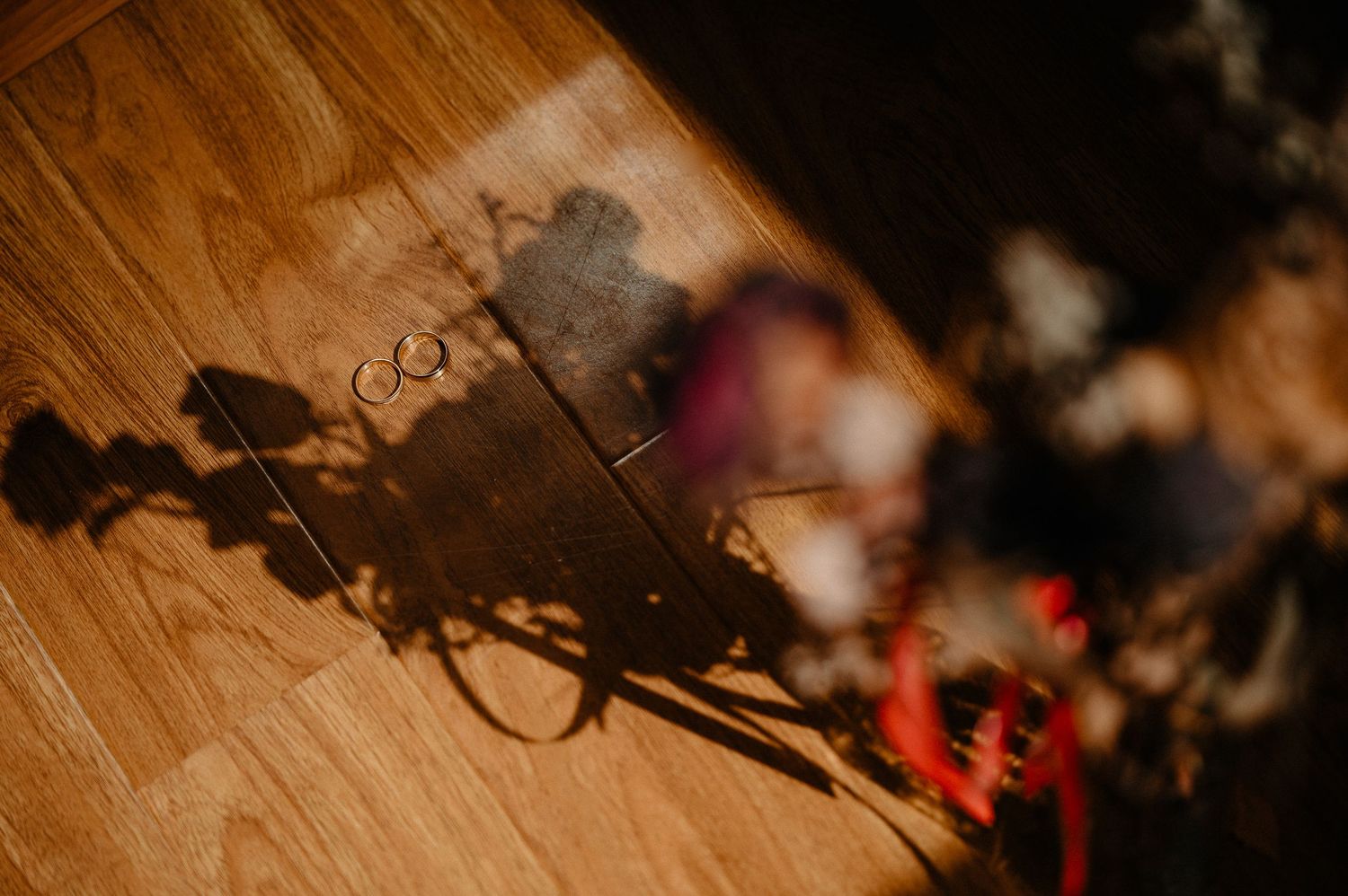 Wedding rings casting shadows on a wooden floor beside blurred floral decorations.