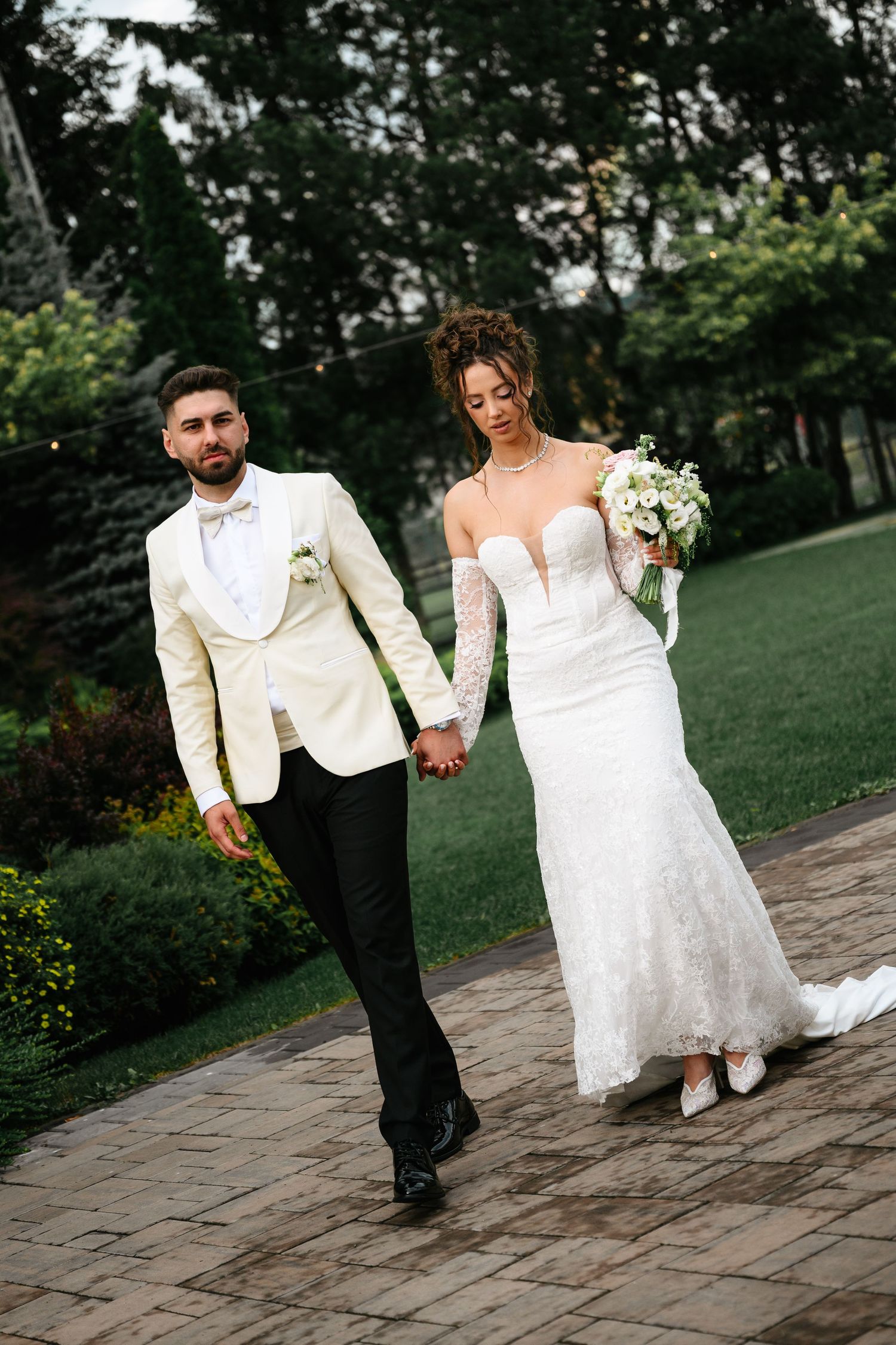 A bride in white lace gown holding bouquet walks hand in hand with groom in ivory tuxedo on garden path.