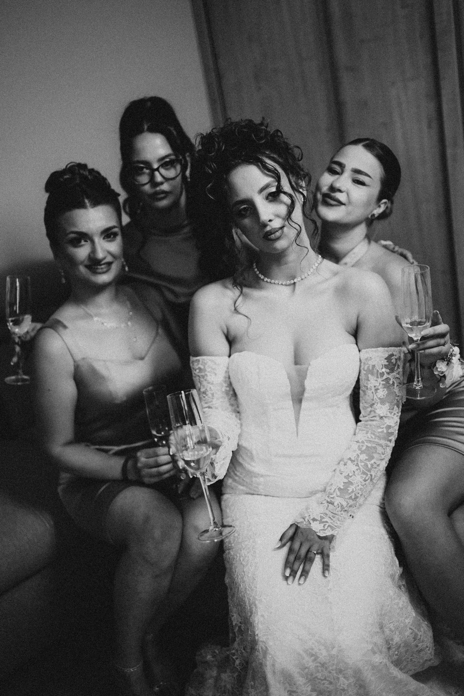 Black and white photo of a bride in white lace gown with bridesmaids holding champagne glasses.