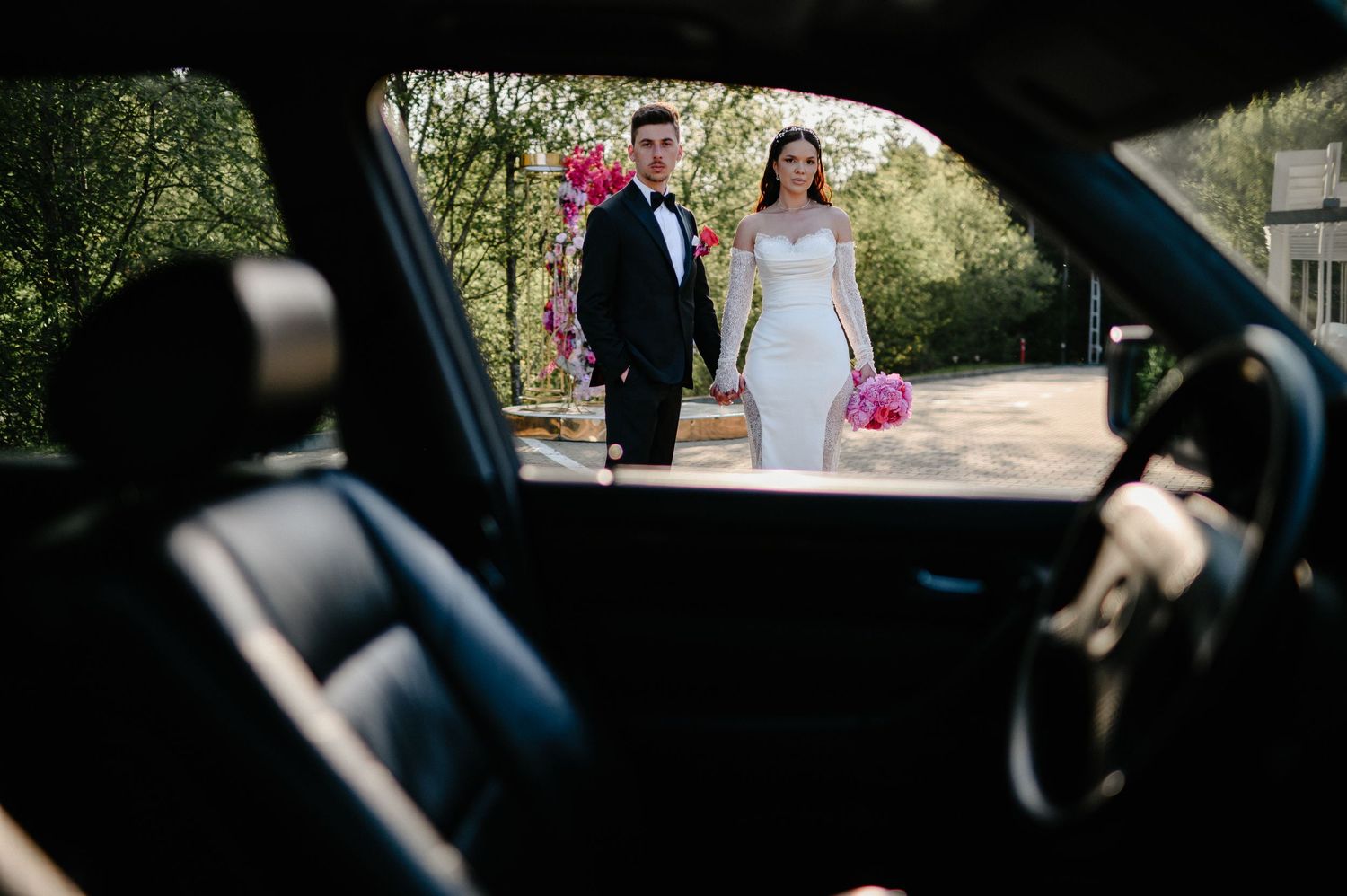 Bride and groom holding hands viewed through vintage car window with pink floral arch background.