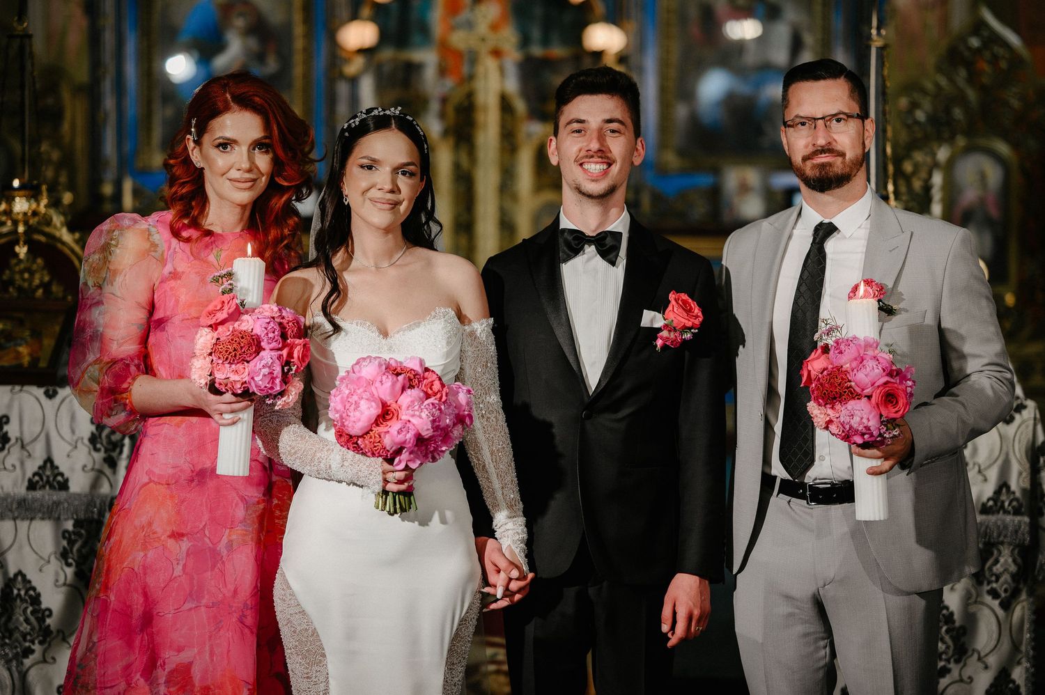 Bride and groom pose with wedding party holding pink floral candles inside an ornate Orthodox church.
