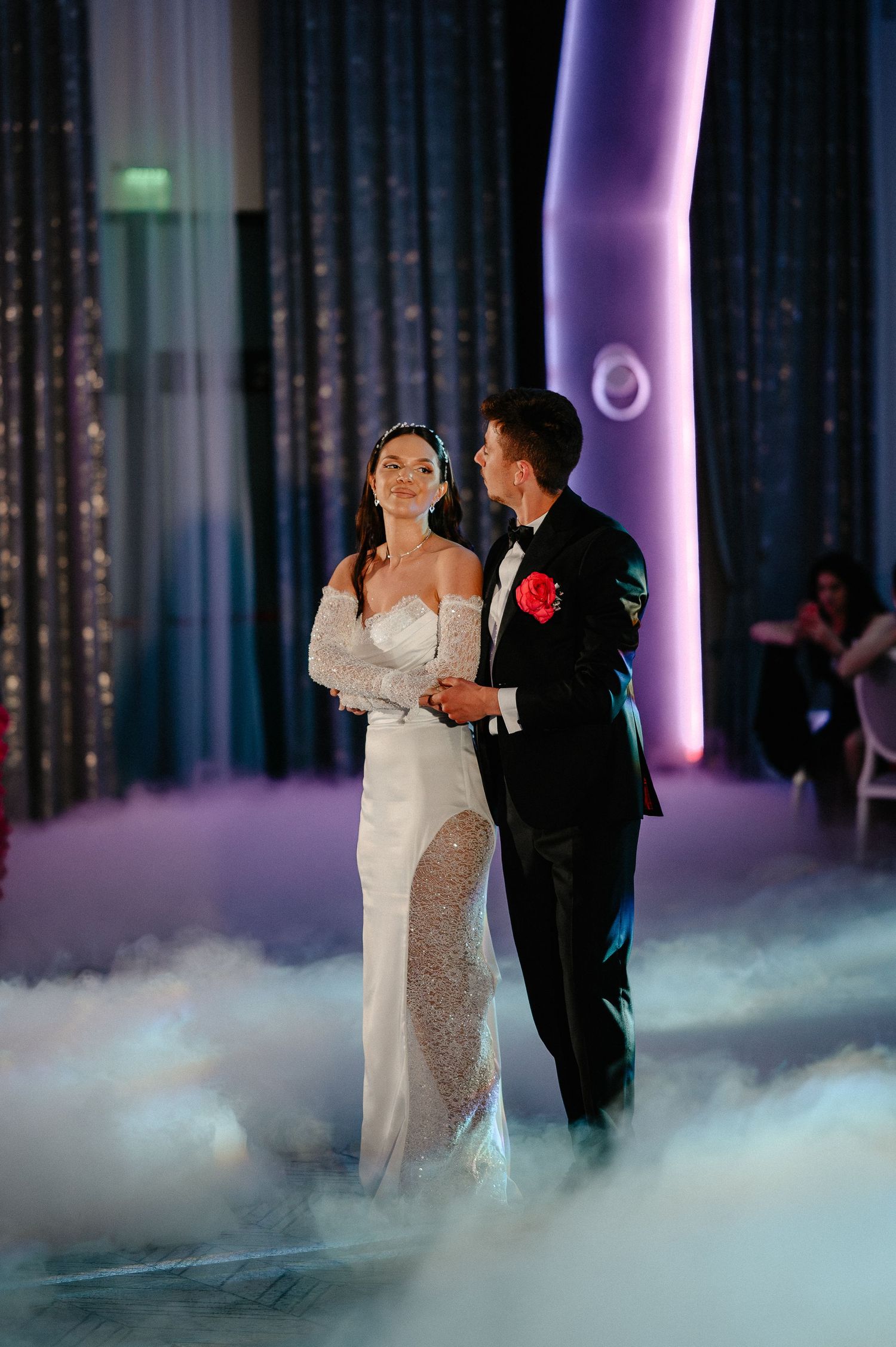 Bride in elegant white gown and groom in tuxedo dance together in dreamy fog at wedding reception.