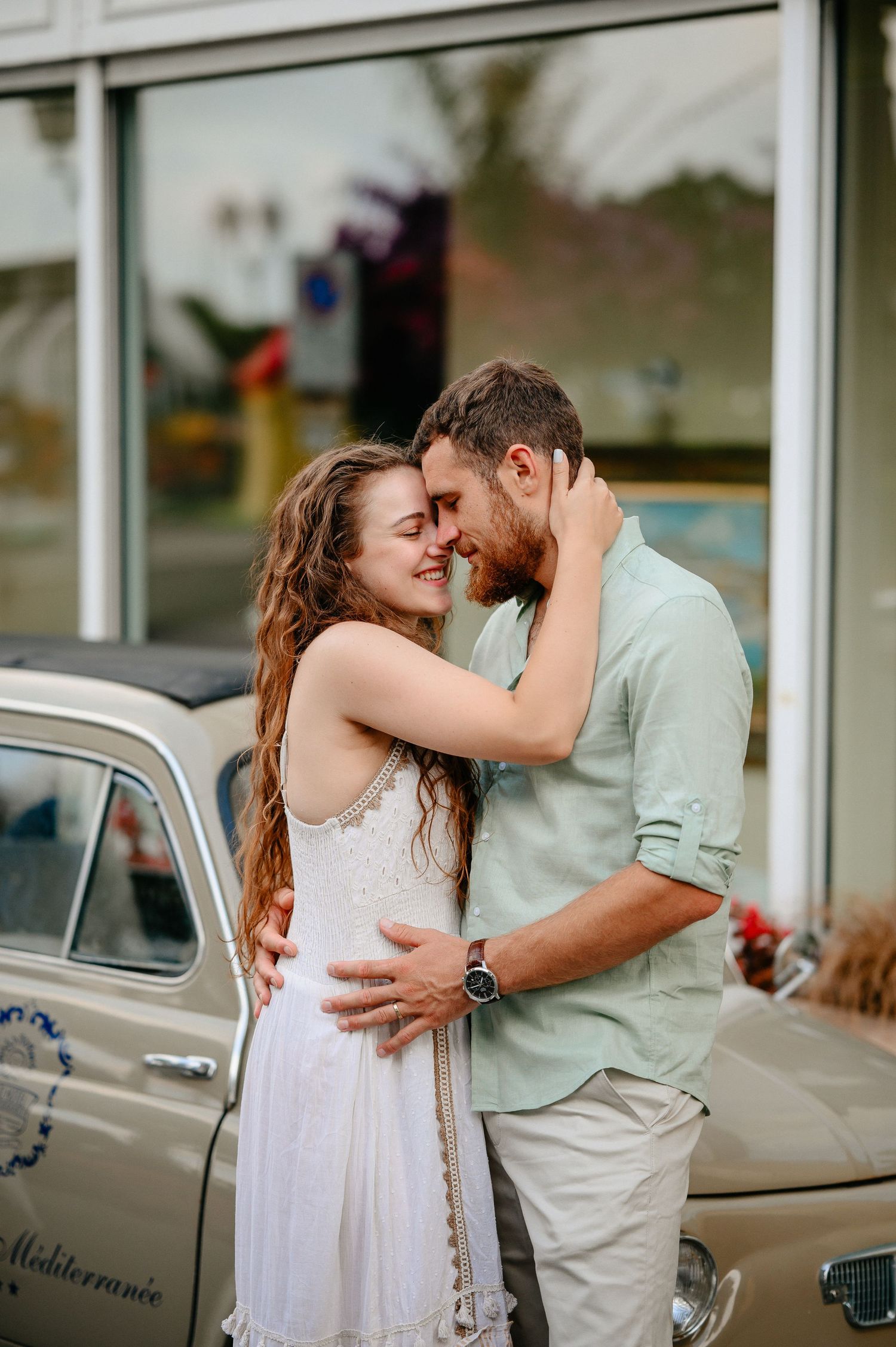 Romantic couple embracing and touching foreheads beside a vintage beige car outside a shop.
