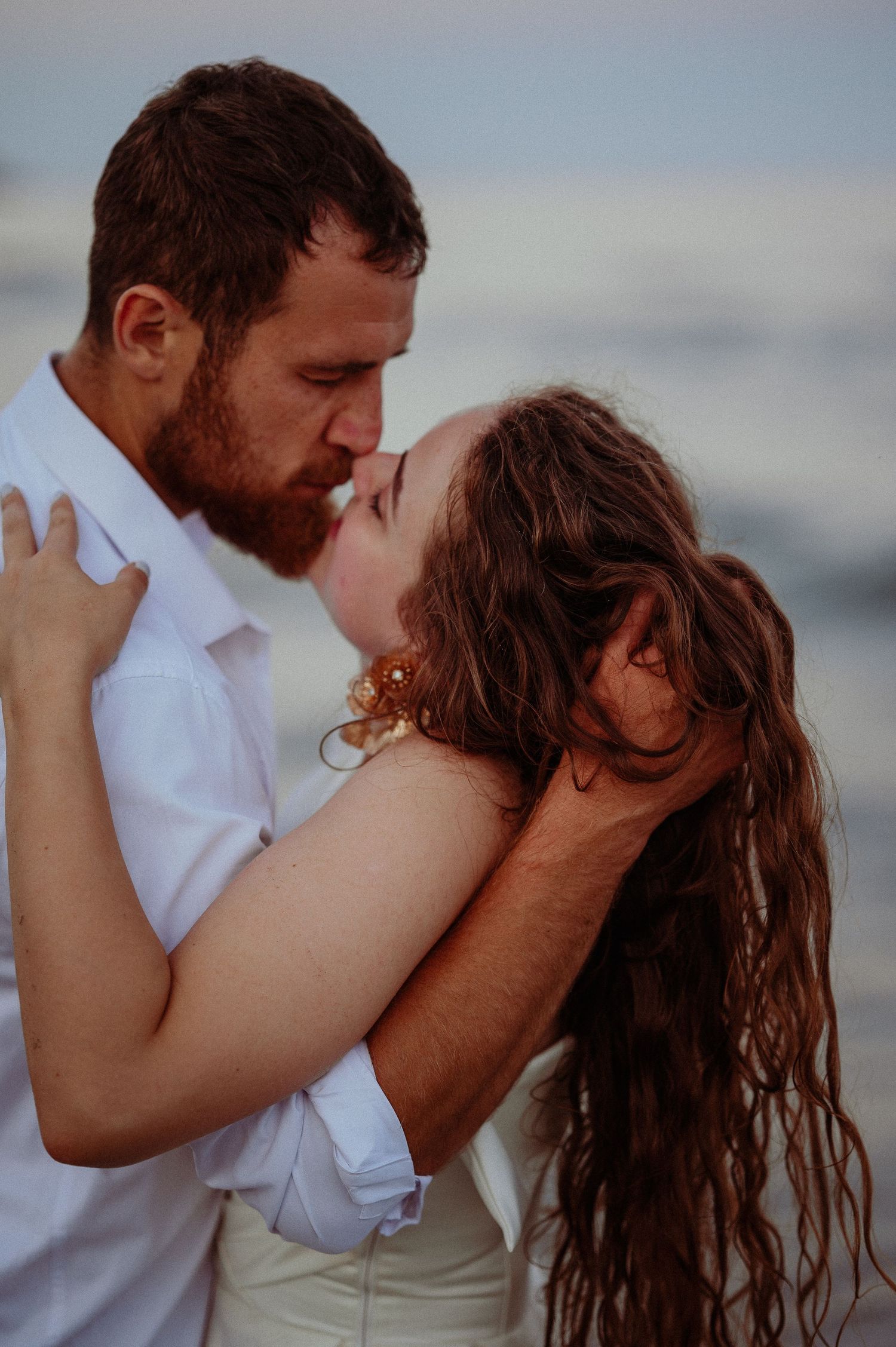 Bearded groom in white shirt kissing bride with long wavy hair against blurred ocean background at sunset.