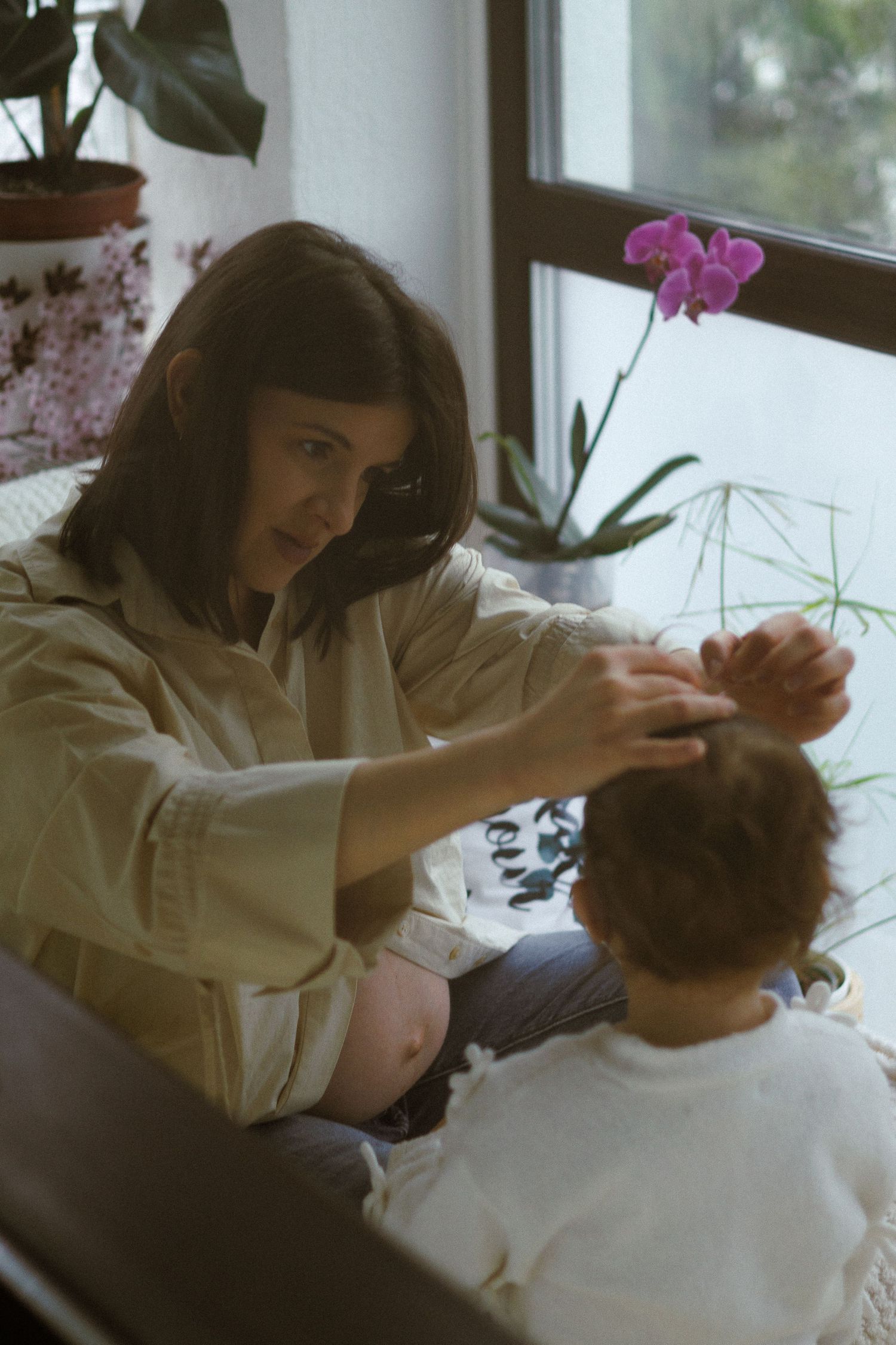 Pregnant woman interacting with young child near window with orchid and houseplants.