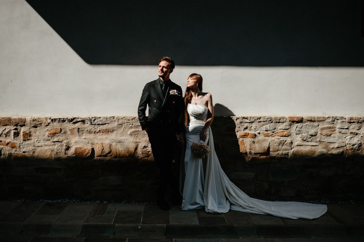 Bride and groom pose against stone wall in striking light and shadow contrast wedding portrait.