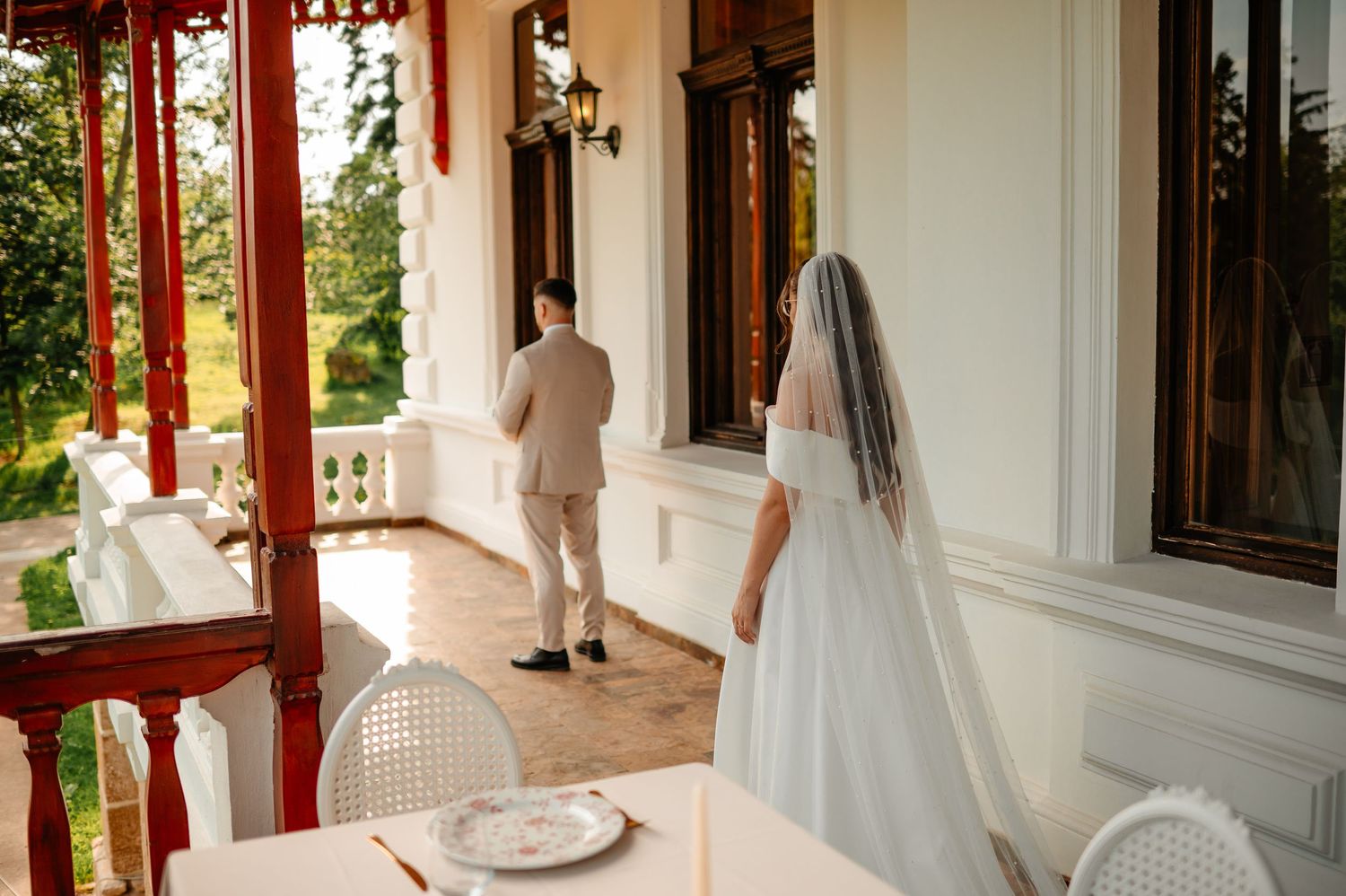 Bride in white gown and veil approaches groom on elegant outdoor veranda surrounded by greenery.