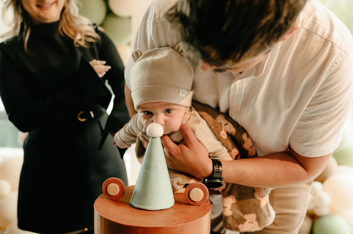 A baby with a pacifier sits near a birthday cake while parents celebrate during a first birthday party.