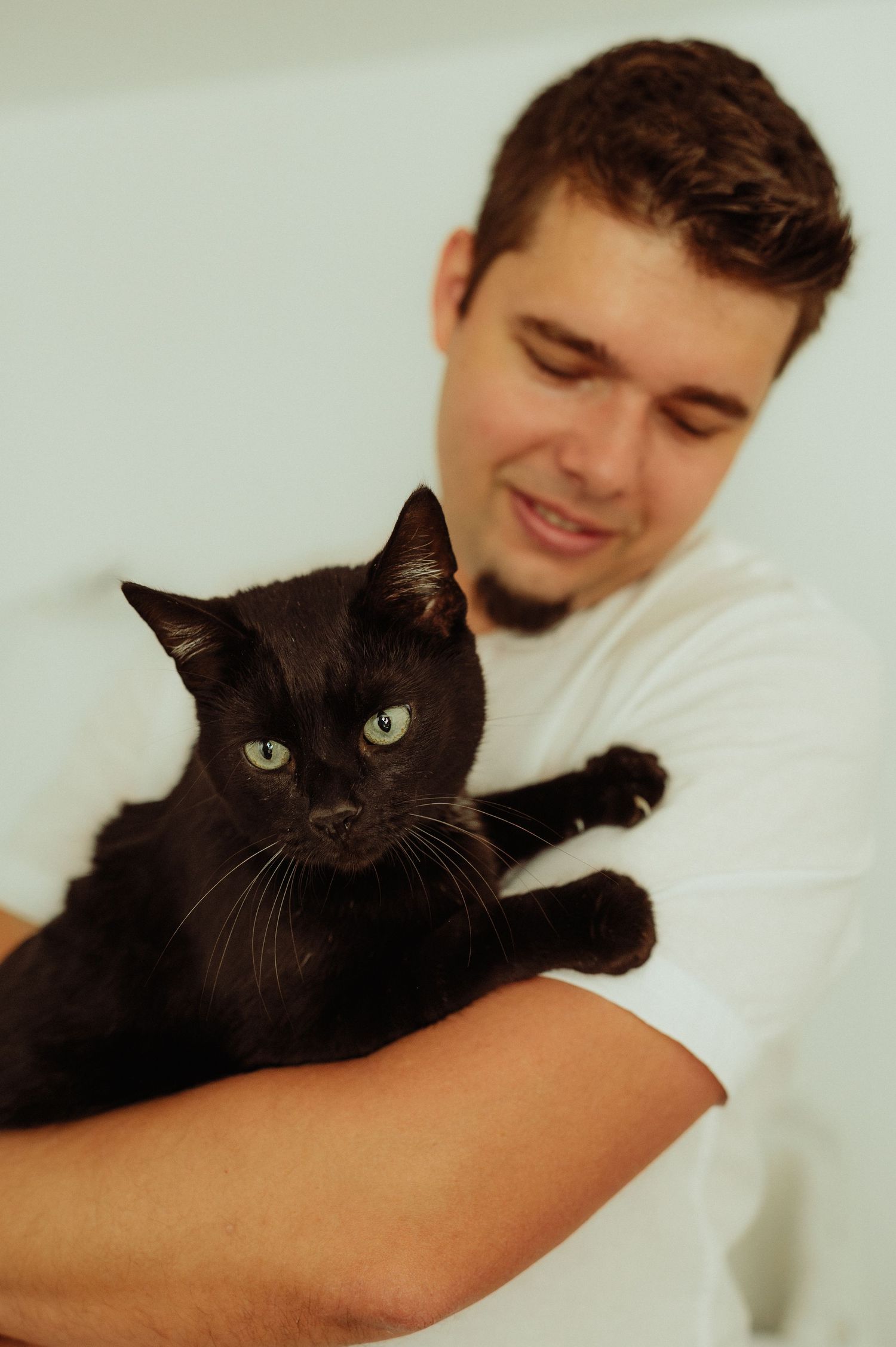 Young man in white shirt smiling while holding black cat with green eyes.