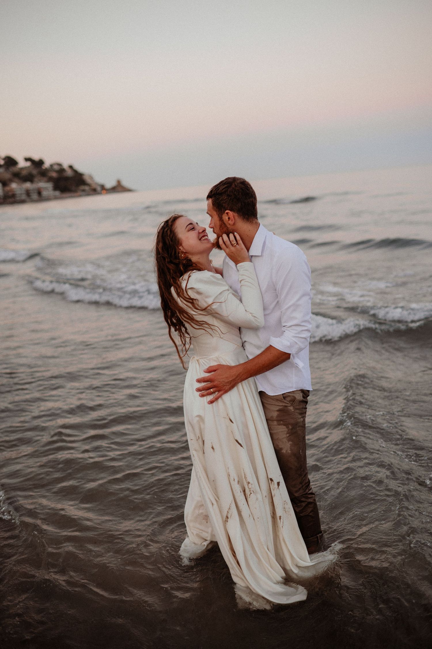 Couple sharing a romantic kiss while standing in ocean waves at sunset, woman in white dress.