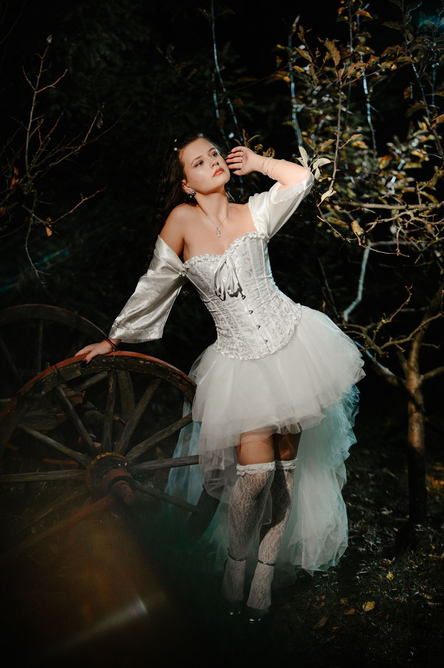 Woman in white corset dress and lace stockings stands beside antique wagon wheel in dark woods.