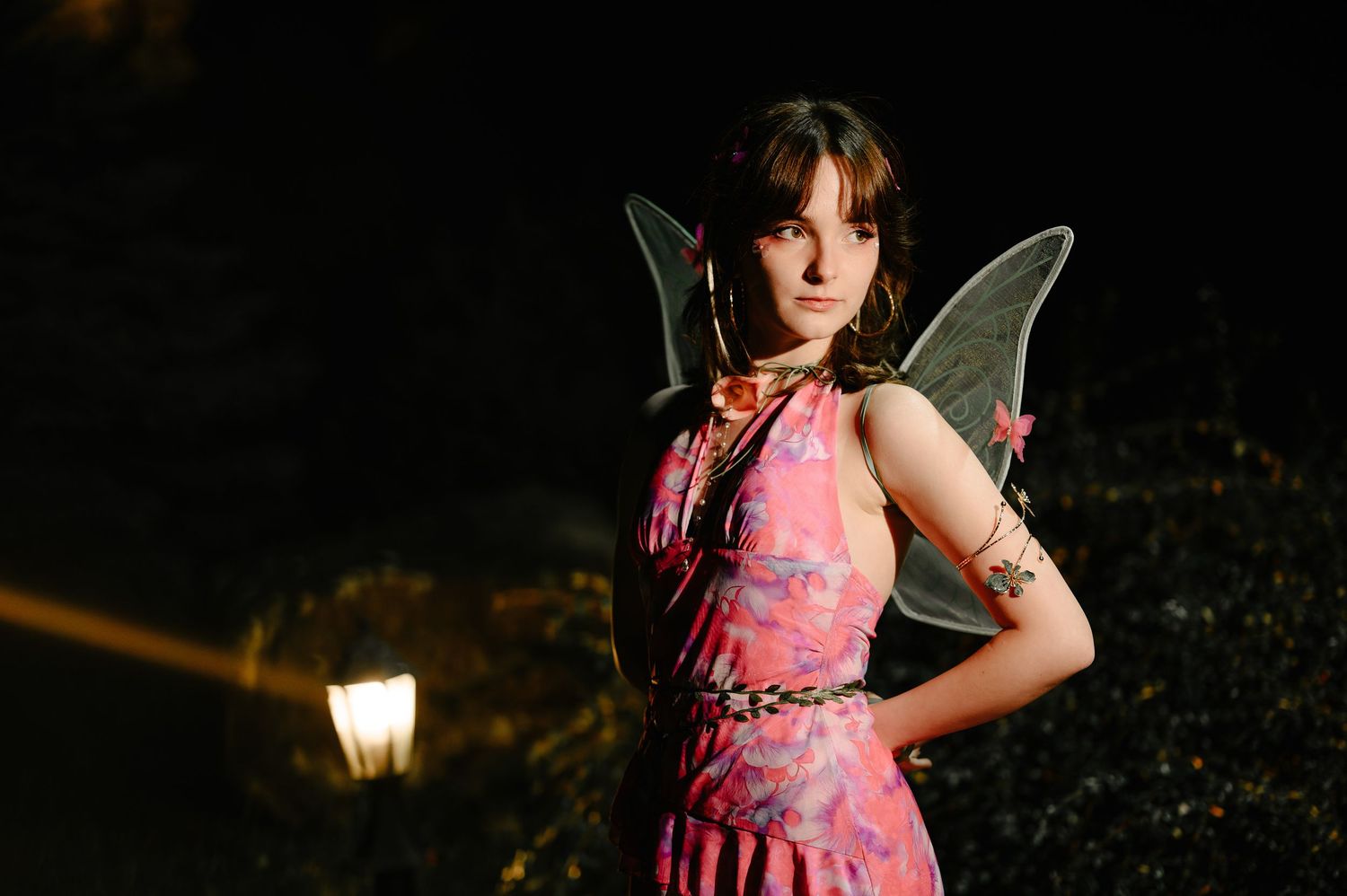 Young woman in pink floral dress with green fairy wings poses near outdoor lamp at night.