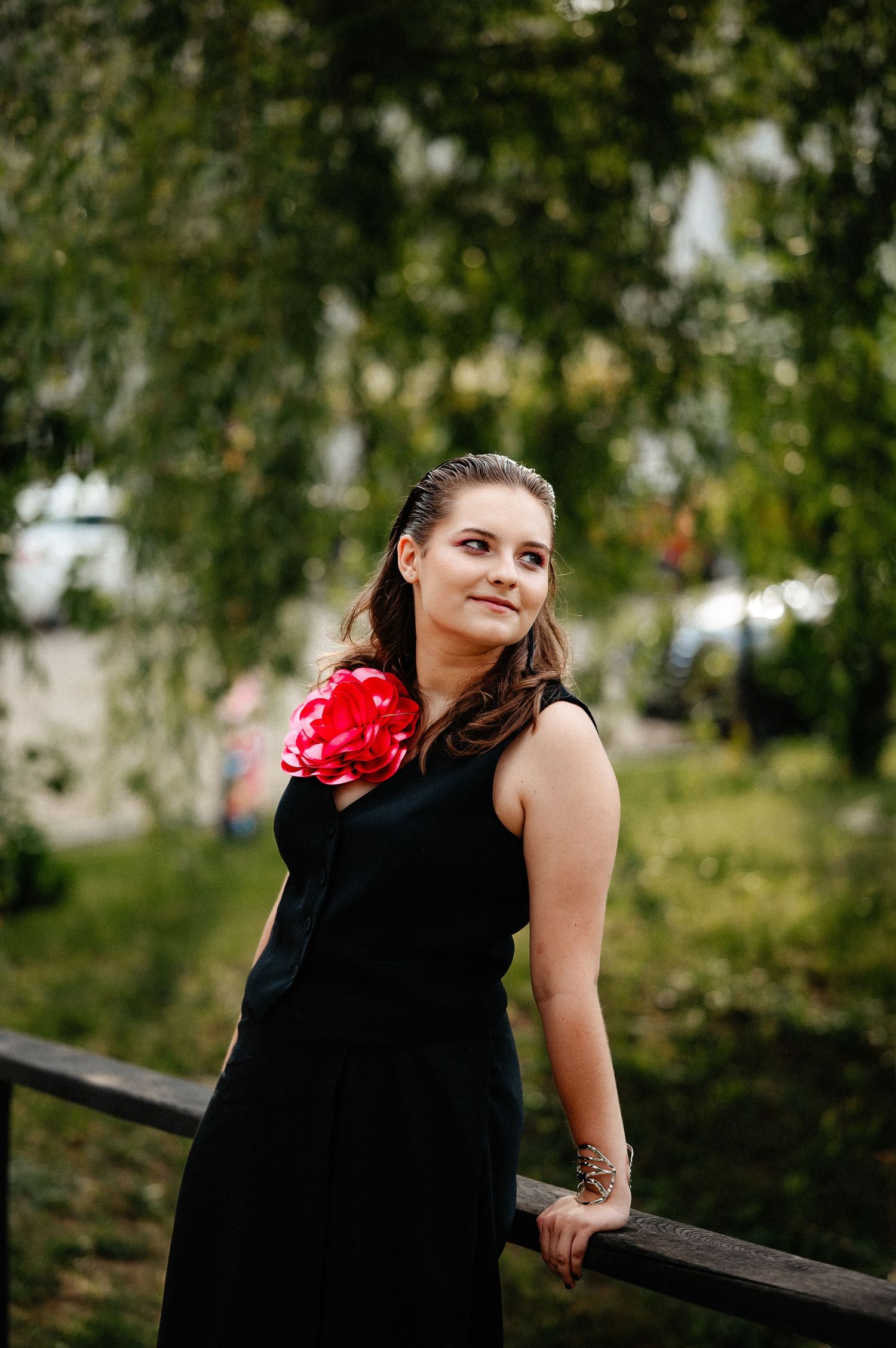 Young woman in black sleeveless dress with red flower corsage leaning on wooden fence outdoors.