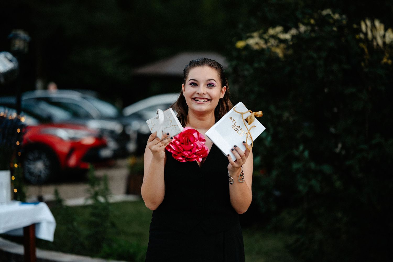 Smiling young woman in black dress holding gifts with red bow at an outdoor party.