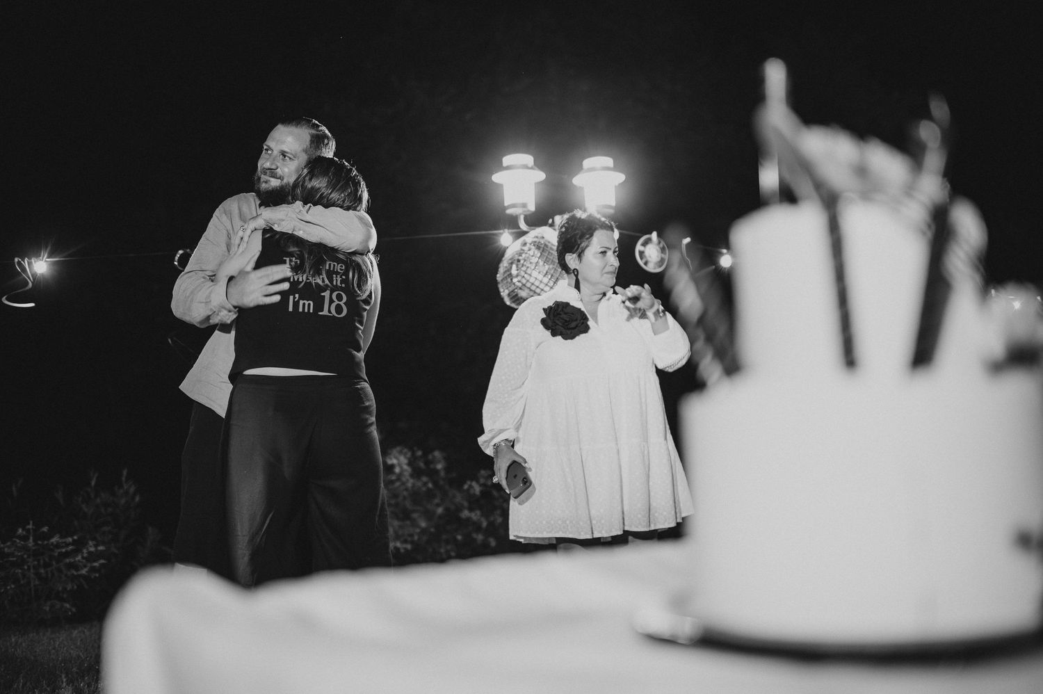 Black and white photo of guests hugging near a birthday cake at an outdoor night event.