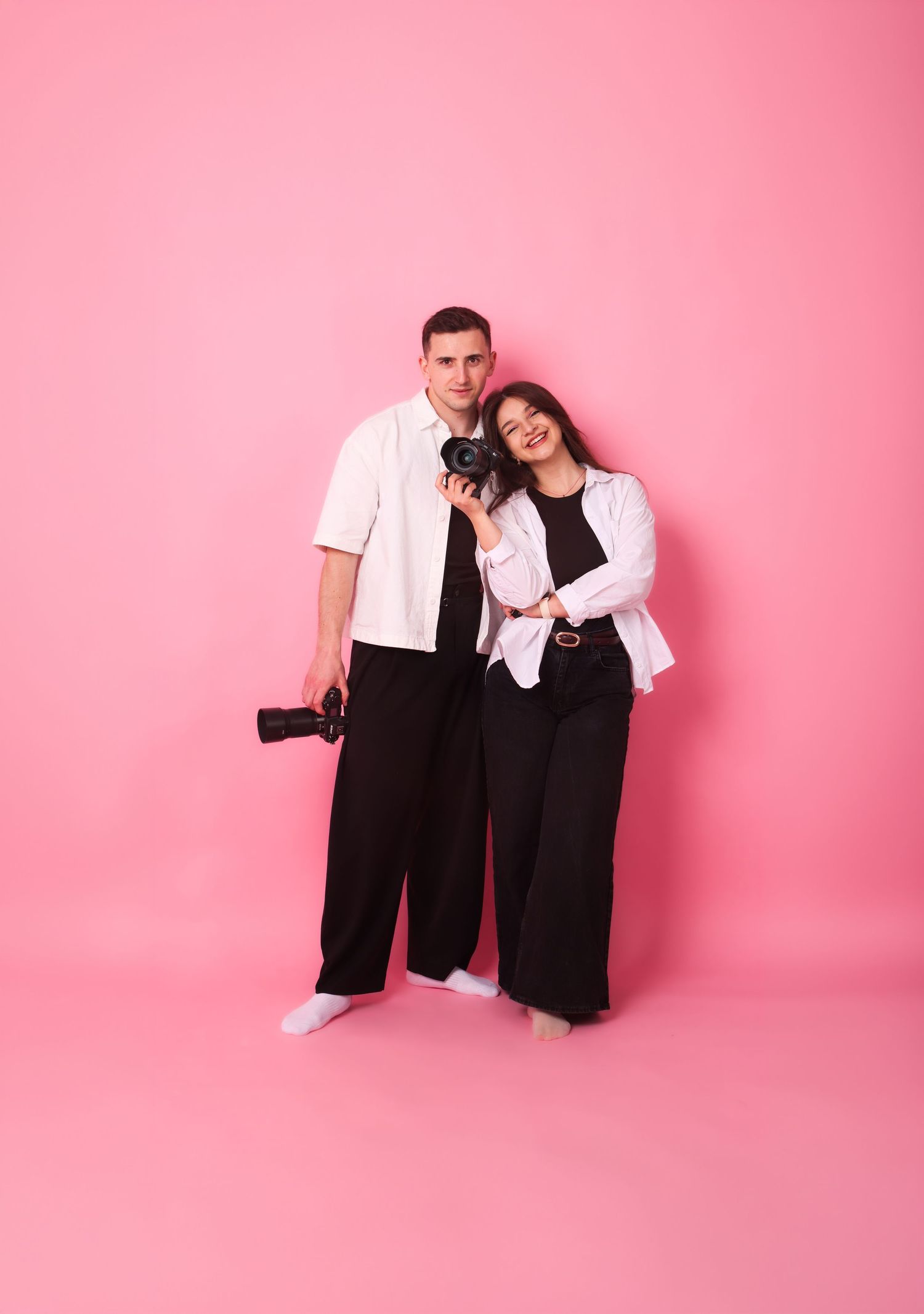 Couple posing against a pink background, both dressed in black and white, holding professional cameras.